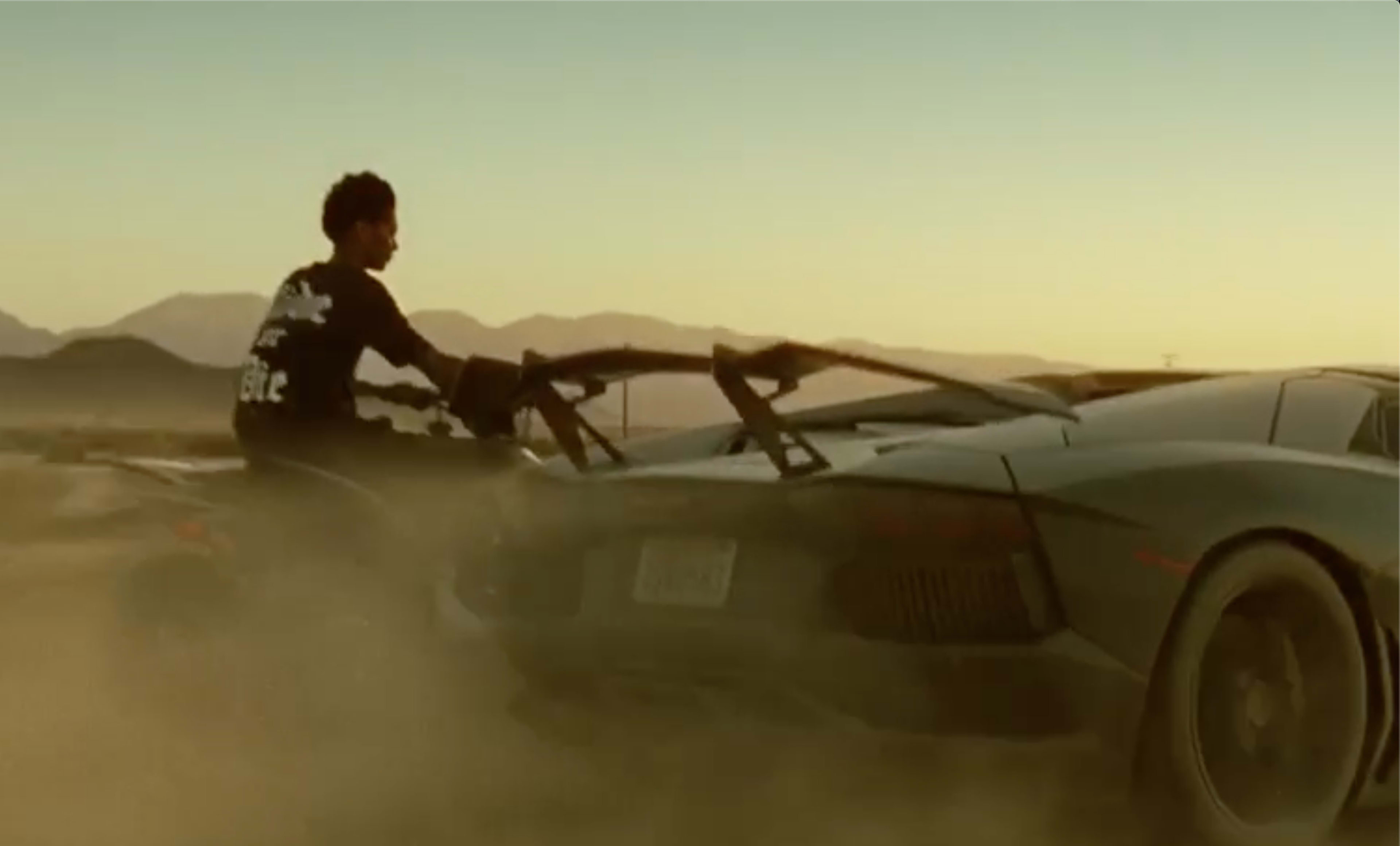A person rides an all-terrain vehicle beside a parked sports car in a dusty landscape during sunset.