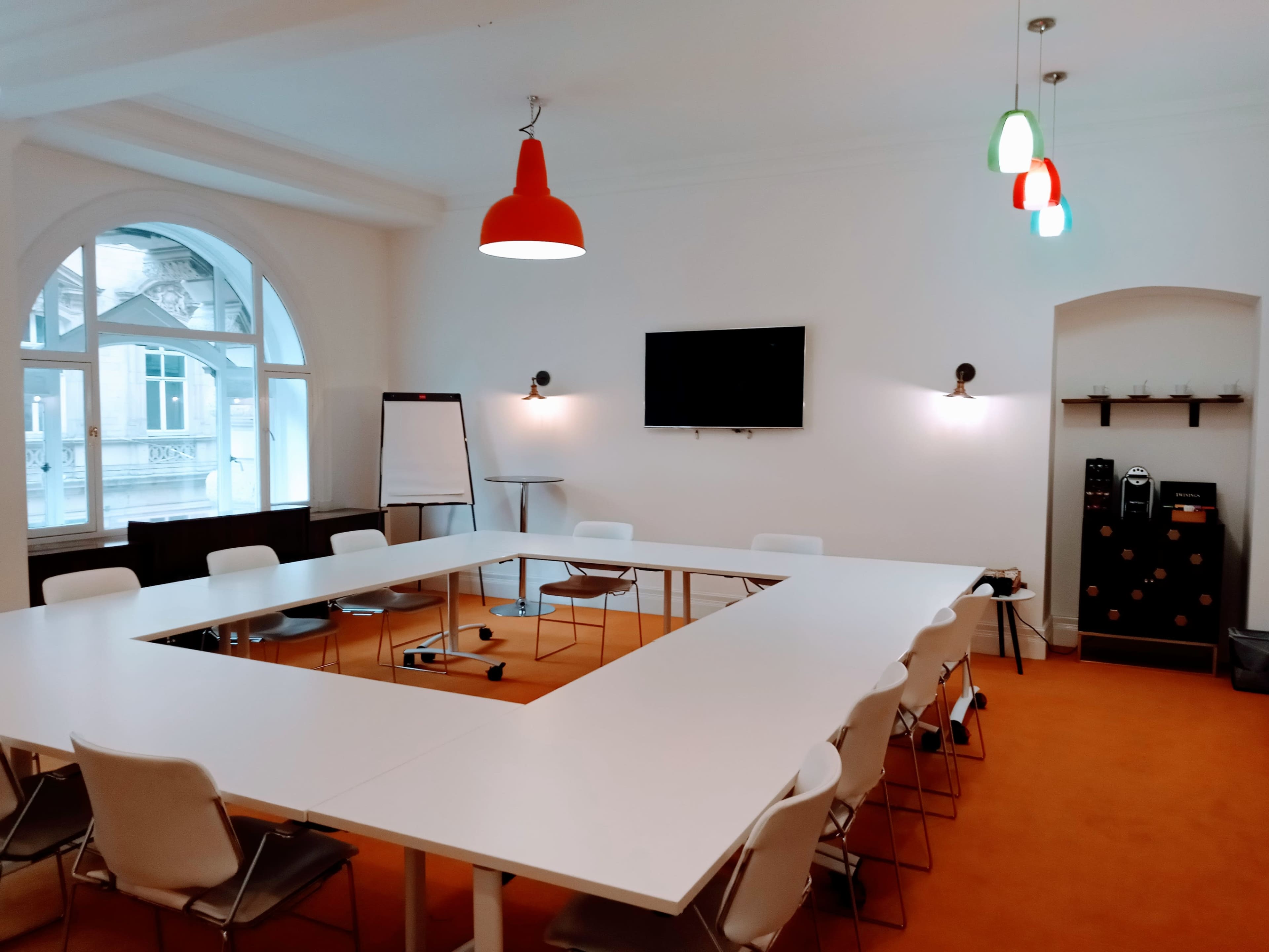 A conference room features a white U-shaped table surrounded by chairs, a television mounted on the wall, and a window letting in natural light.