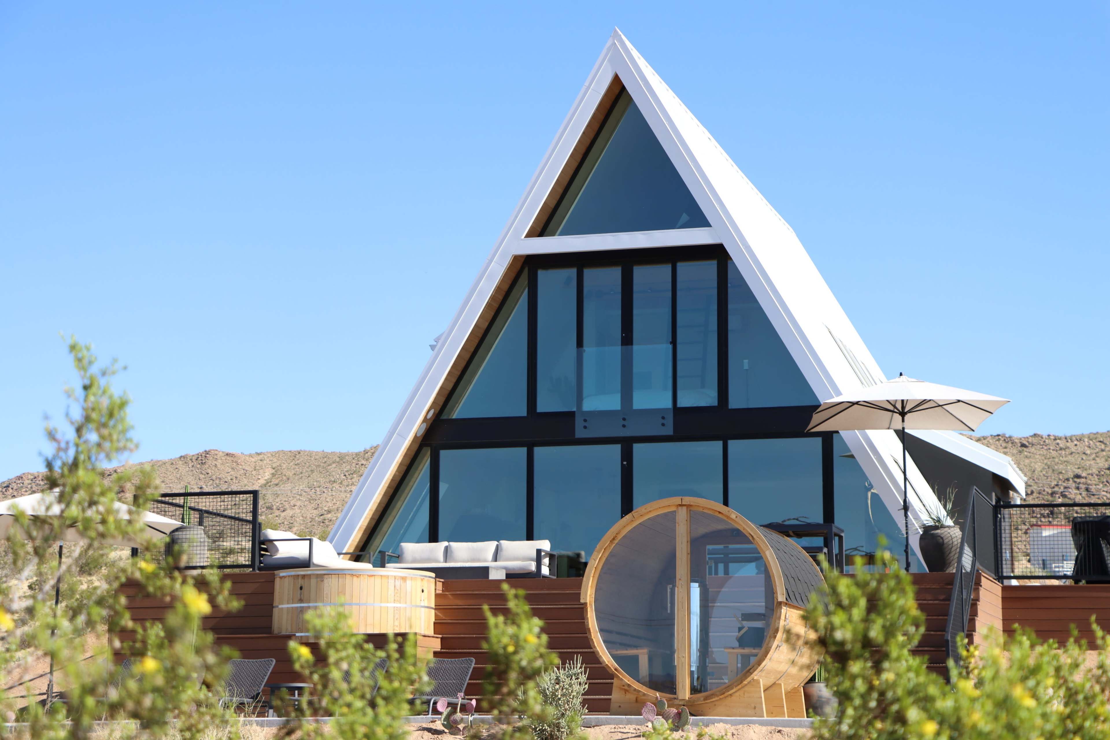 A modern A-frame house with large windows is set against a clear blue sky, featuring a wooden deck and a round, wooden hot tub nearby.