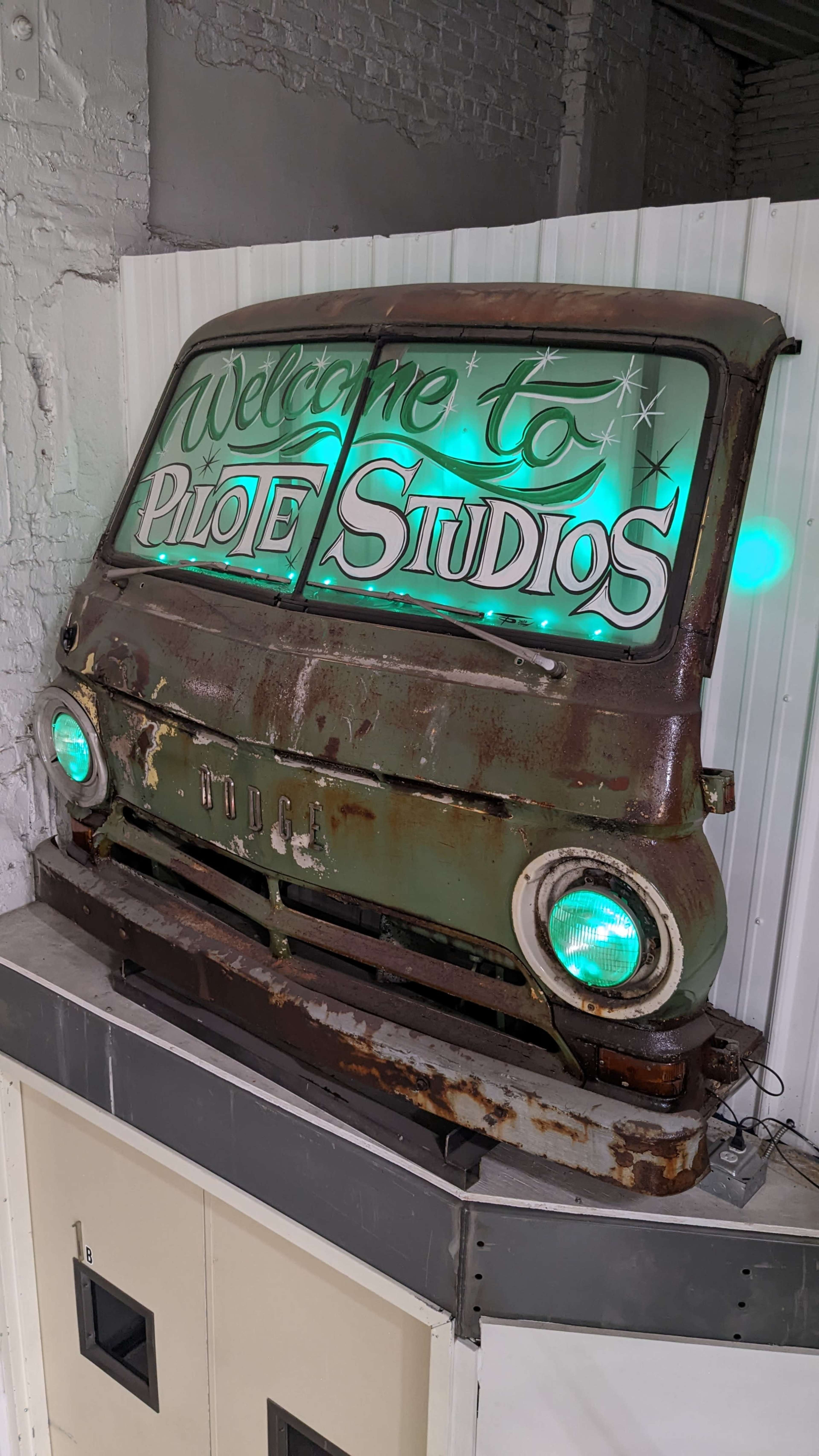 A vintage, rusted vehicle front is mounted on a wall, featuring illuminated text that reads "Welcome to Pilot Studios."