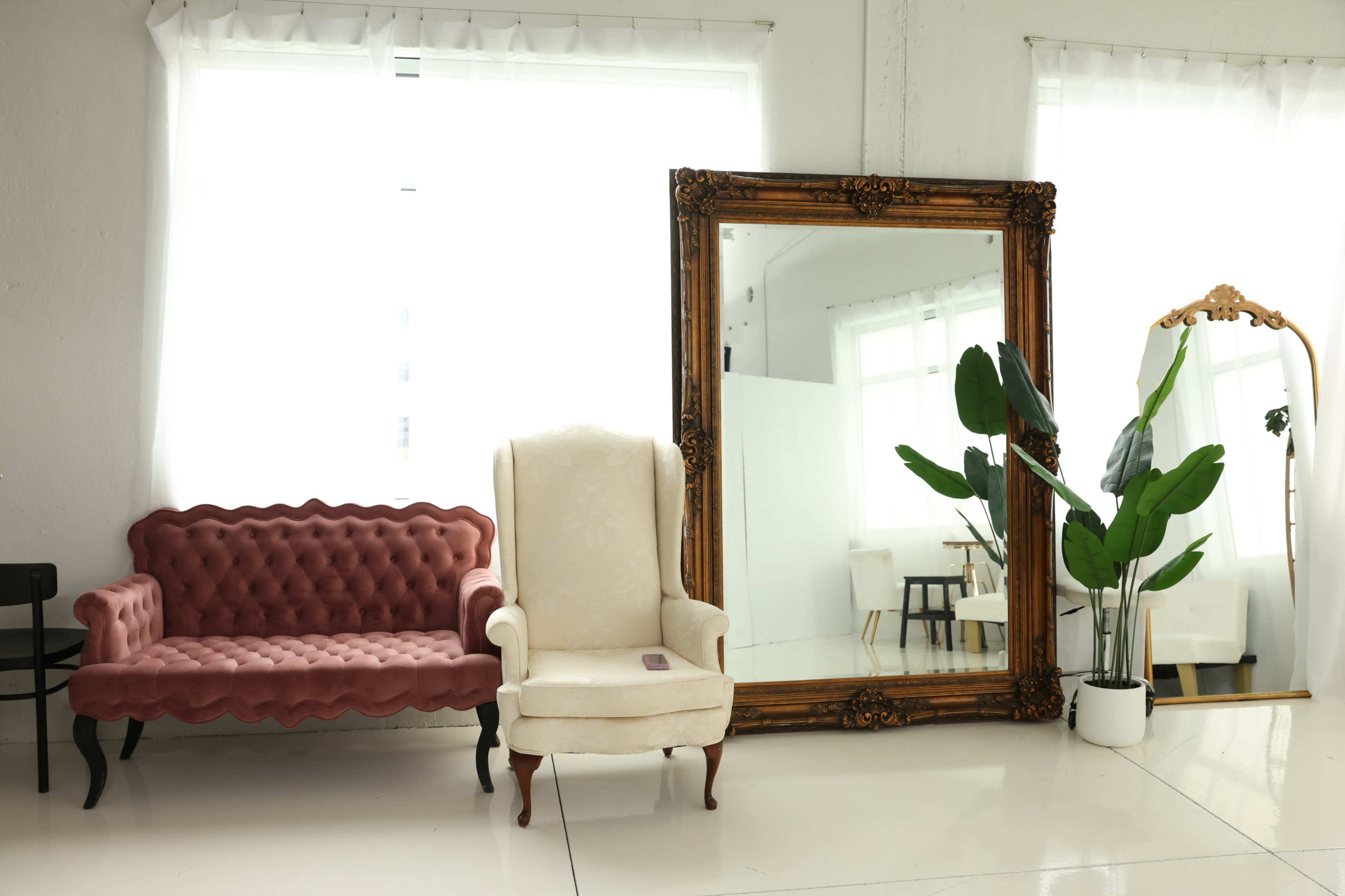 A pink tufted sofa and a cream armchair are positioned beside a large ornate mirror in a well-lit room with white curtains.