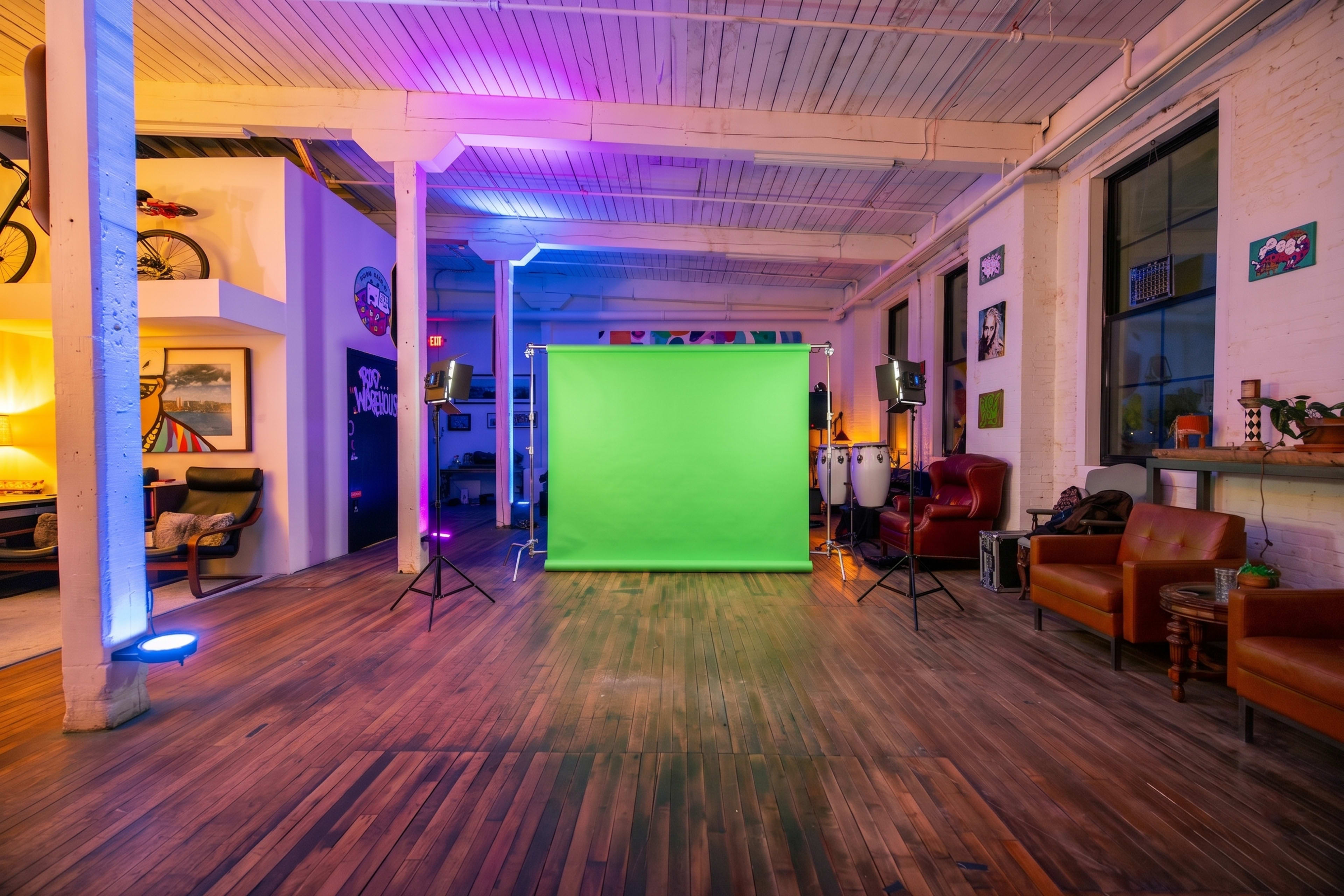 The image shows a spacious studio with wooden flooring, featuring a green screen set up in the center, surrounded by lighting equipment and lounge seating.