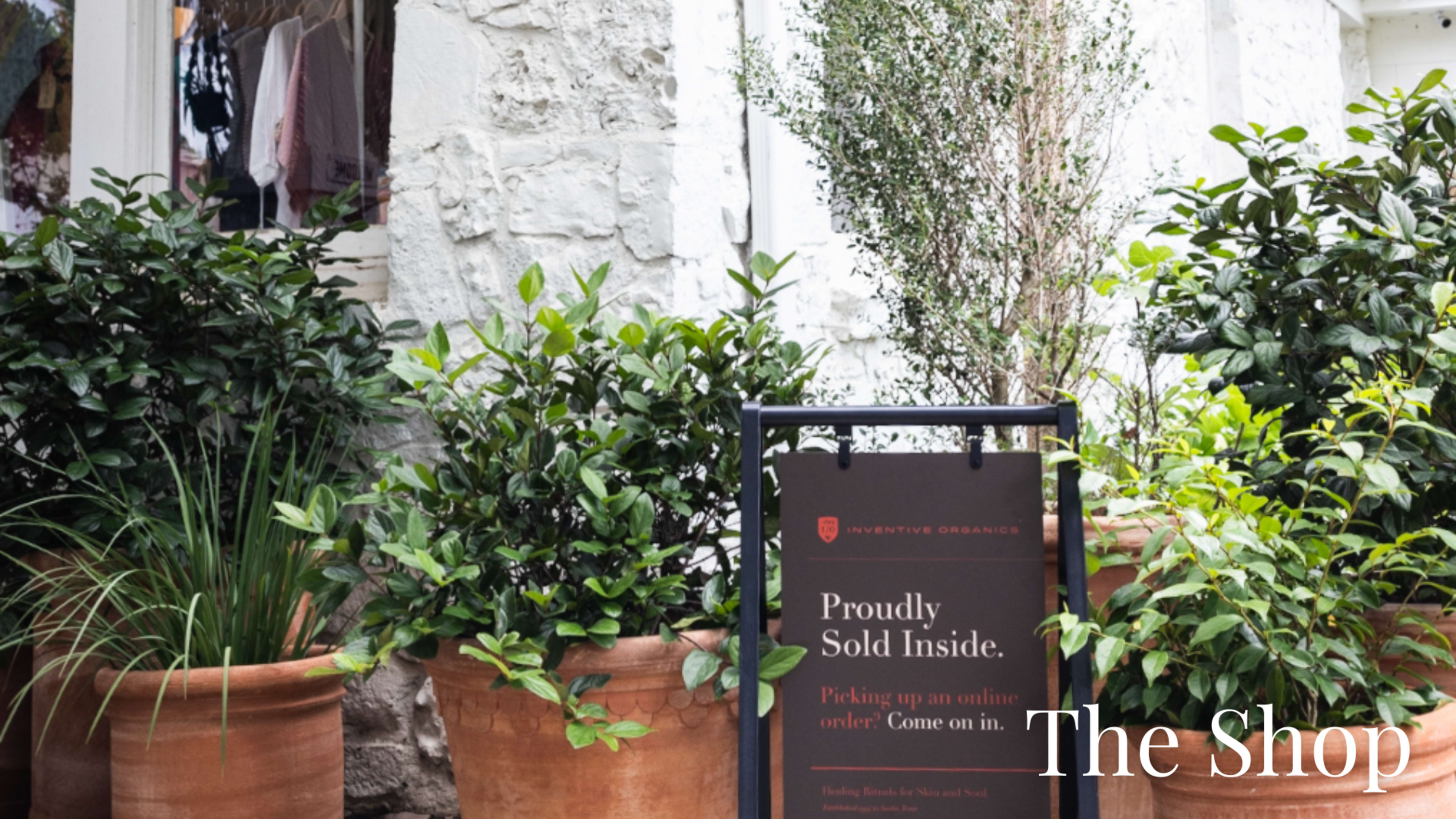 The image shows a sign outside a shop indicating that products are sold inside, accompanied by potted plants on either side.
