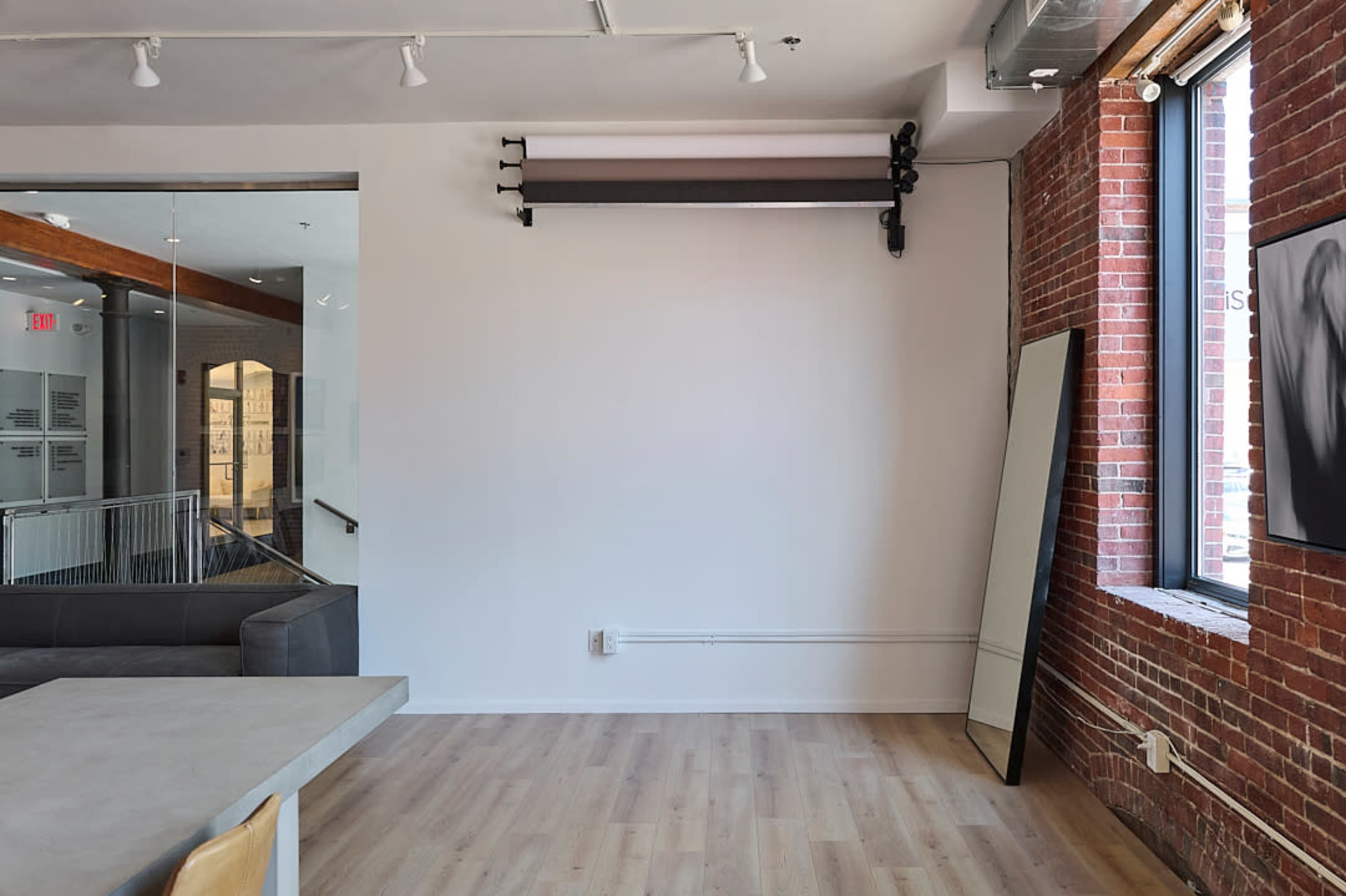 The image shows a minimalist interior of a room featuring exposed brick walls, a large mirror leaning against the wall, and a simple seating area.