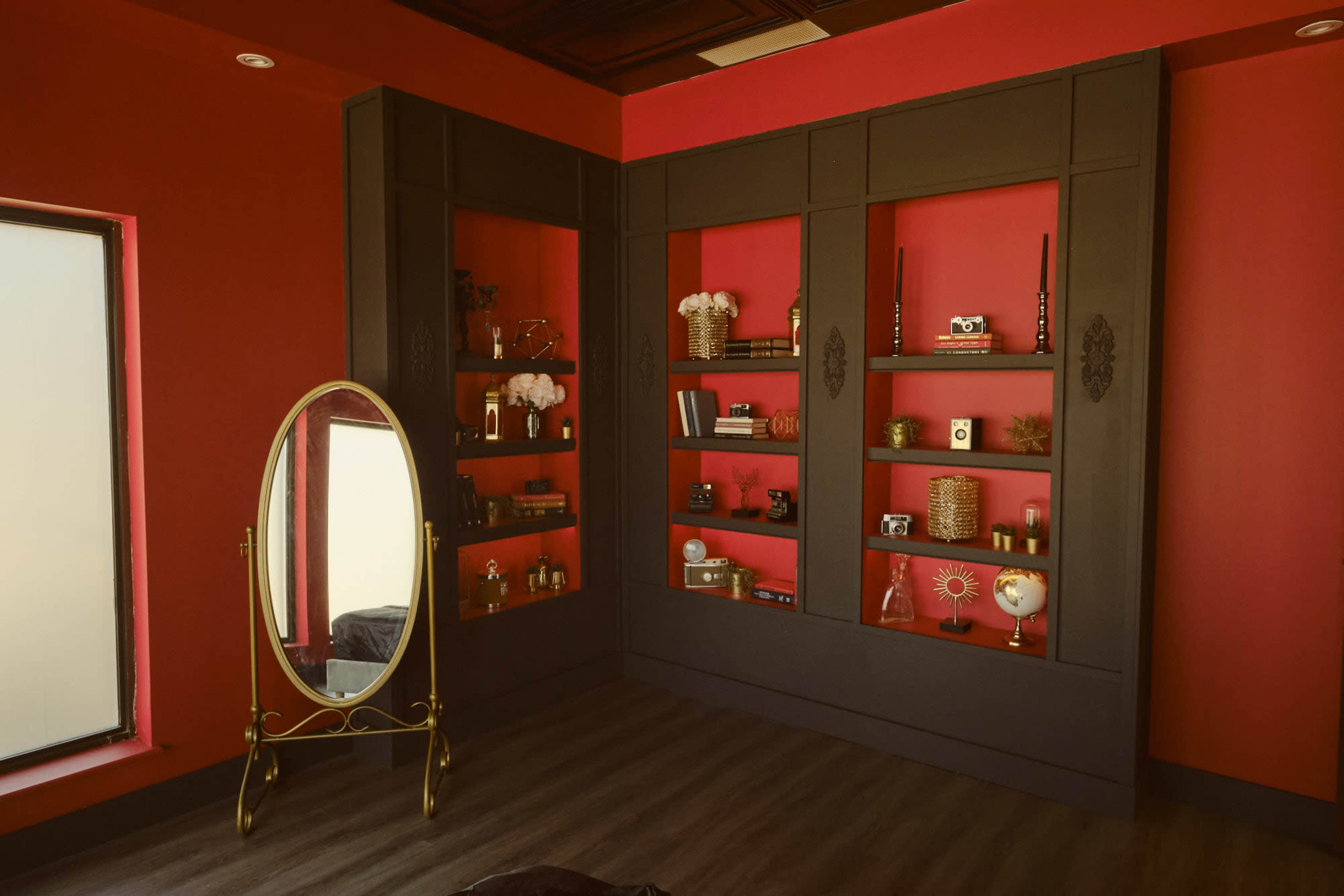 The image shows a corner of a room with dark wooden shelving units, a round mirror, and decorative items against a red wall.