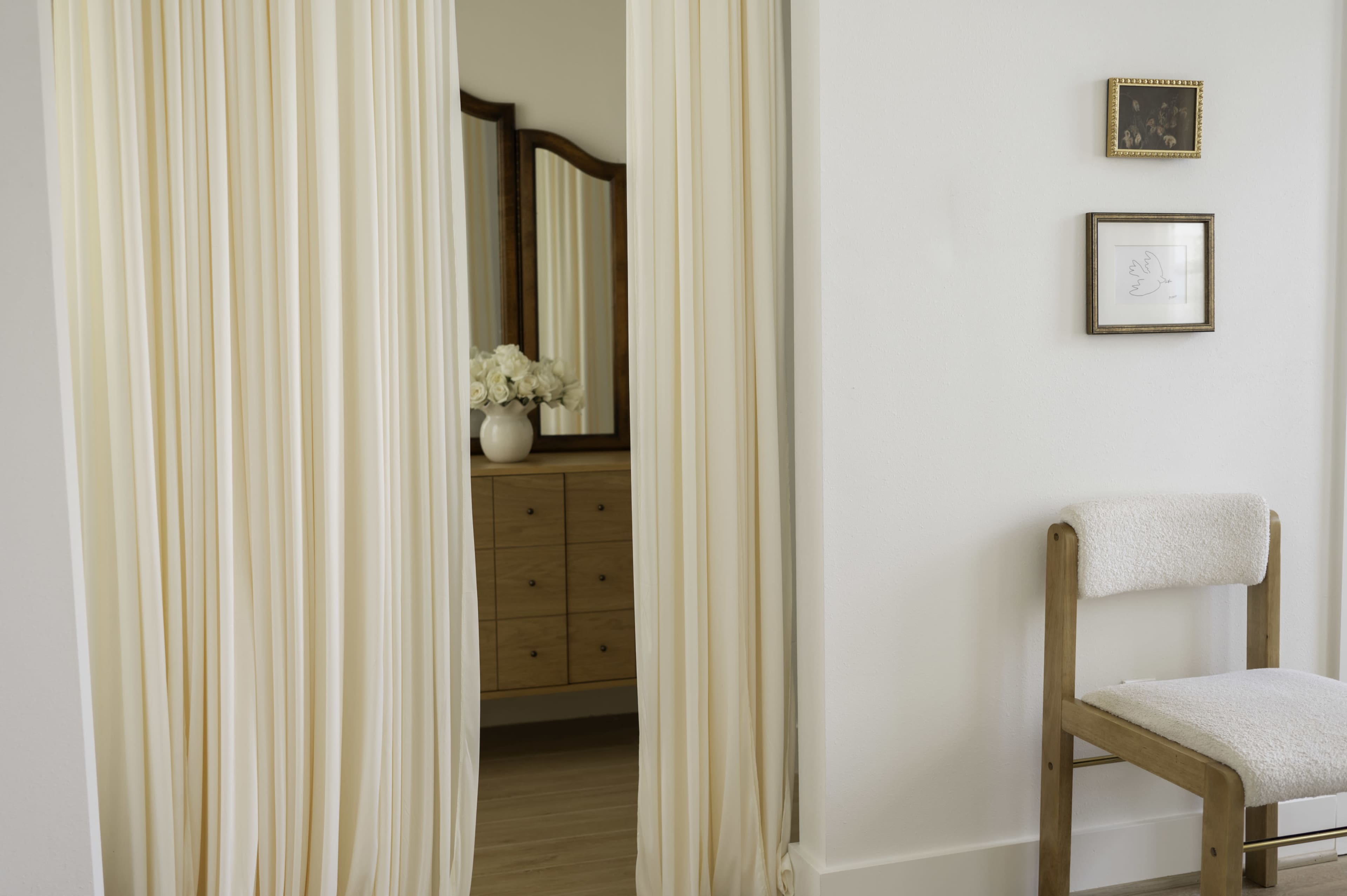 A softly lit room features sheer curtains separating a decorative area with a wooden dresser and a mirror, complemented by a chair on the side.