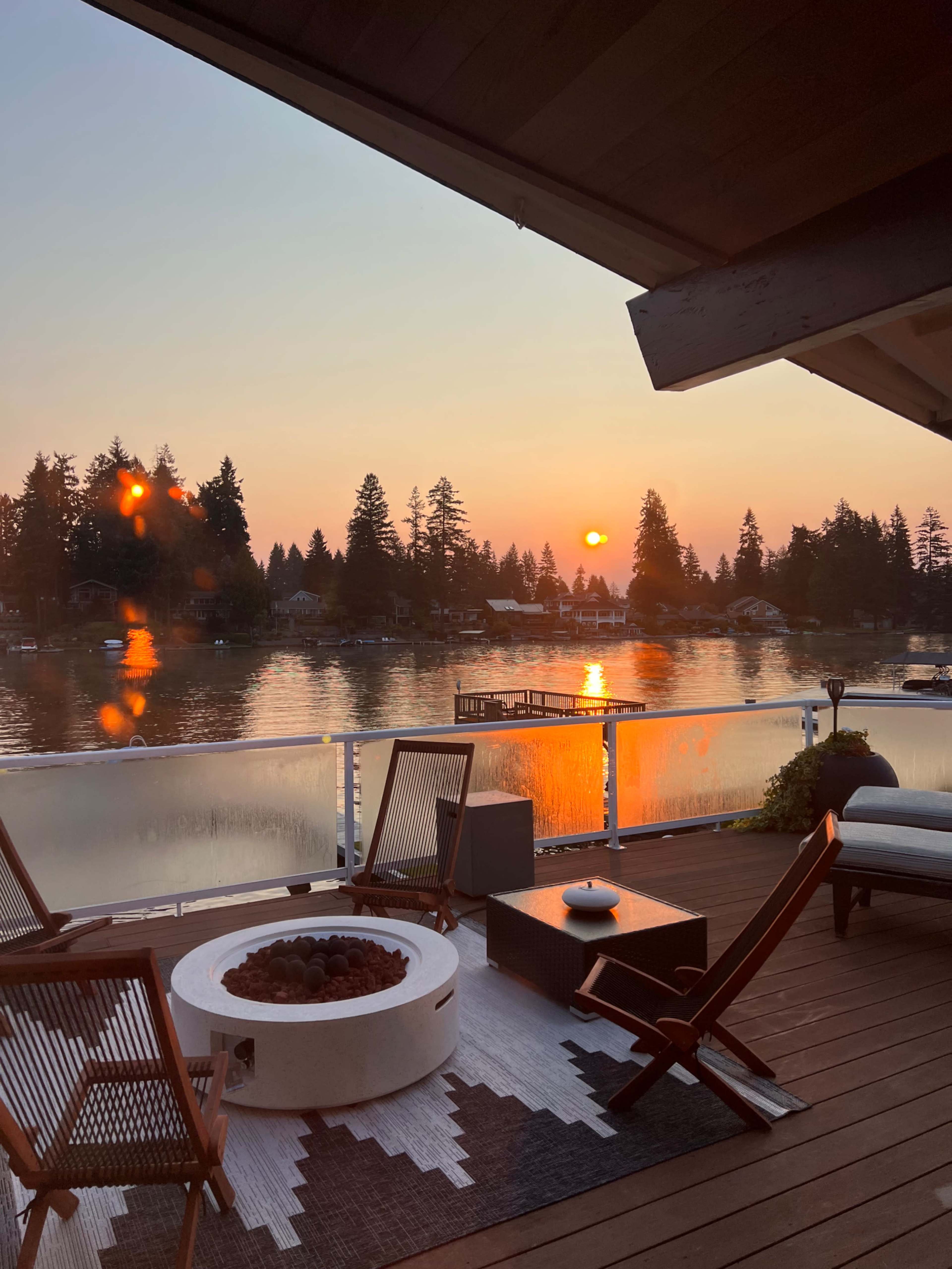 A lakeside deck setup with a fire pit, wooden chairs, and a view of the sunset reflecting on the water.