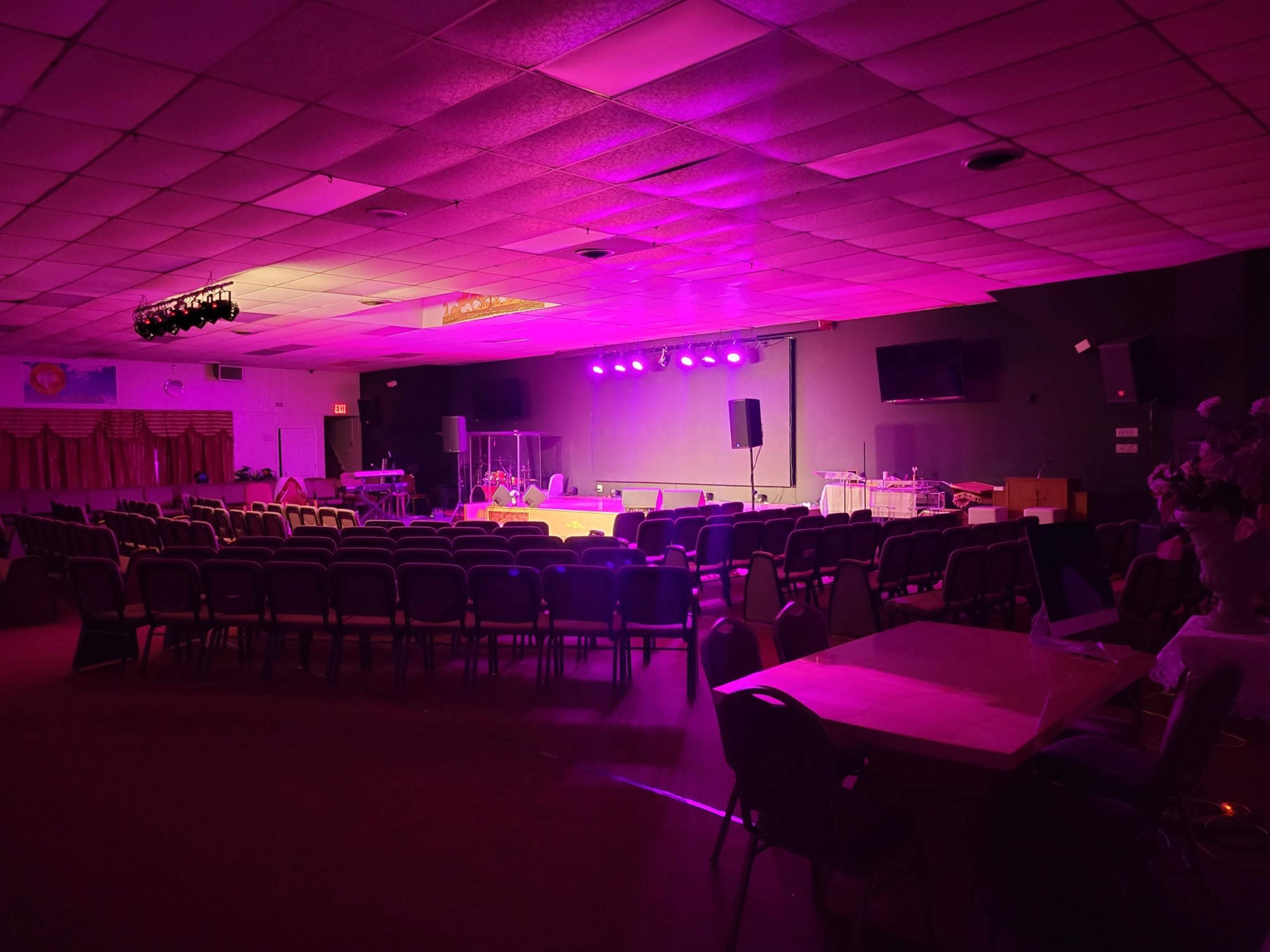 A dimly lit auditorium with rows of chairs facing a stage, illuminated by purple and white stage lights.