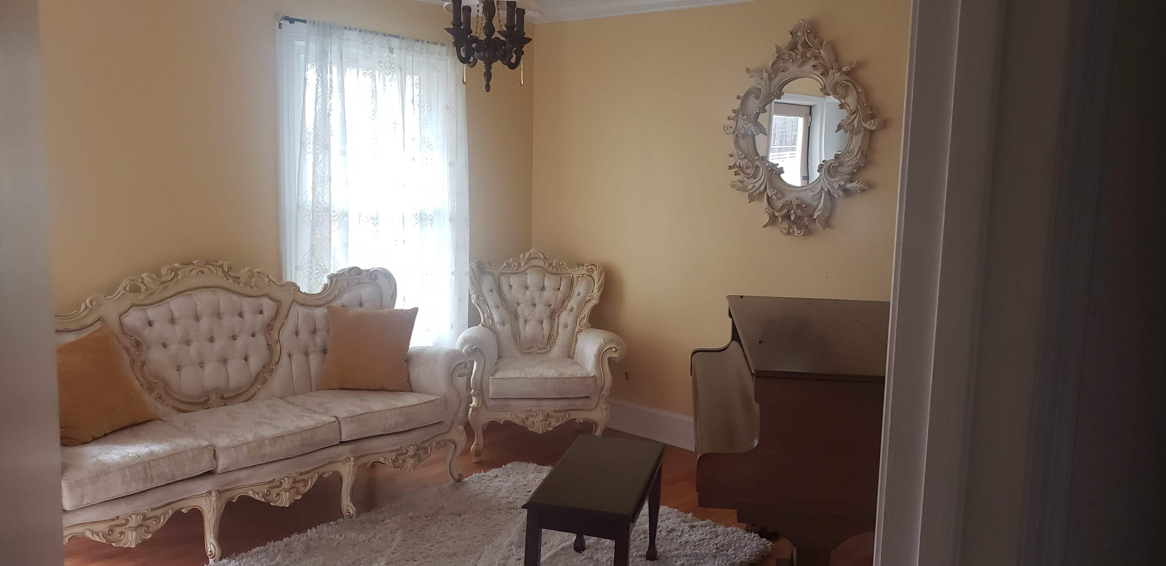 A light yellow-walled room featuring two ornate chairs, a small black table, a piano, and a decorative mirror on the wall.
