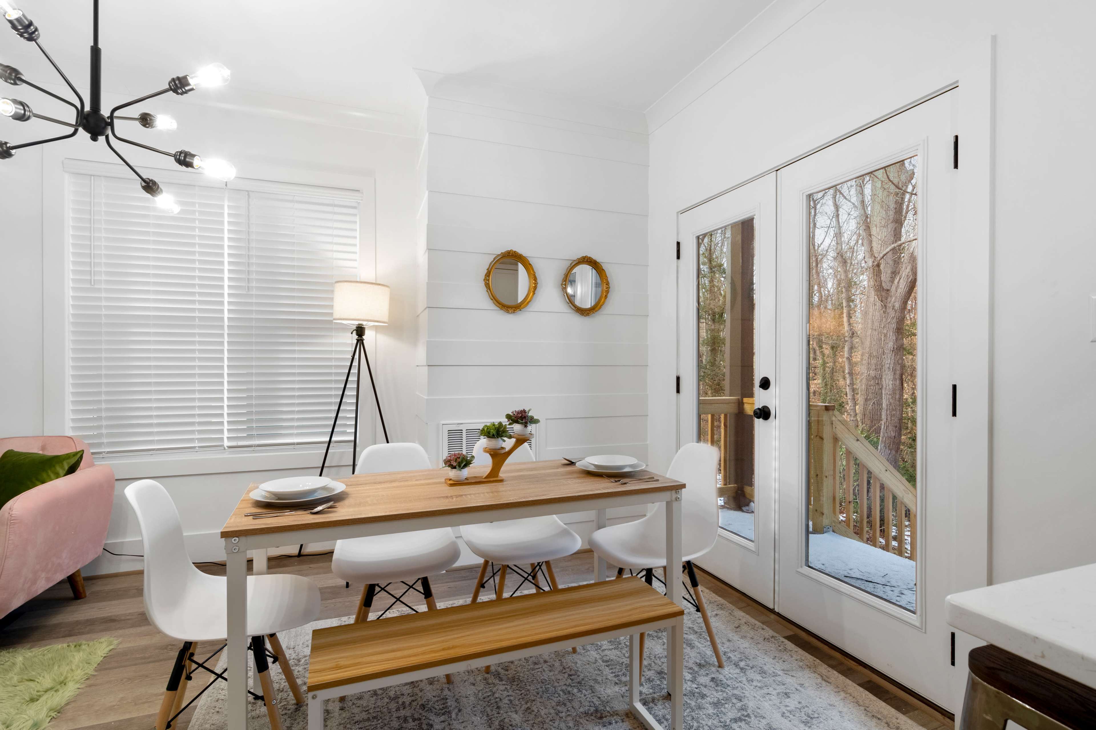A bright dining area features a wooden table with white chairs, a floor lamp, and double glass doors leading to an outdoor deck.