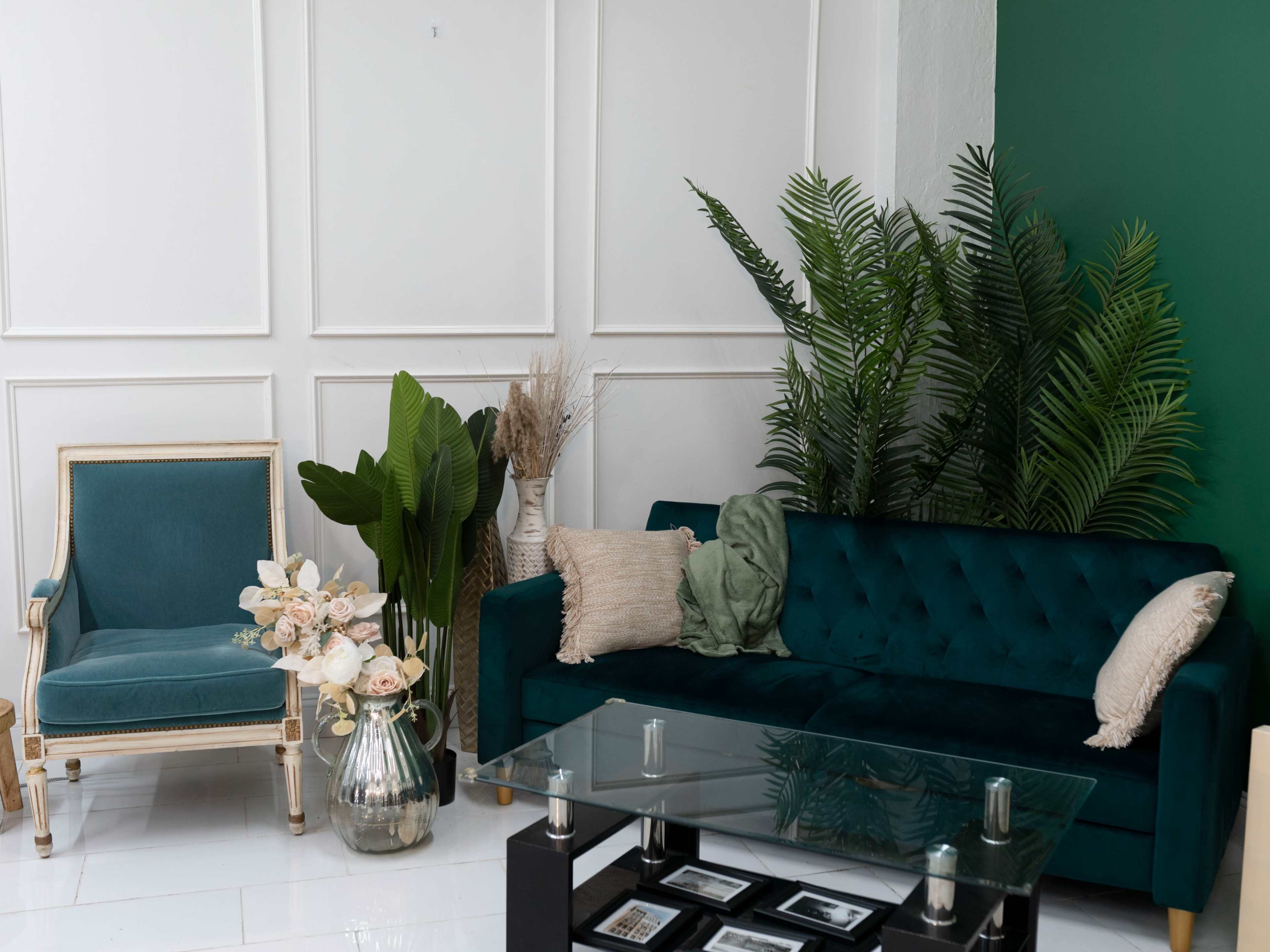 A modern living space features a teal velvet sofa and a vintage-style chair with decorative plants and a glass coffee table.