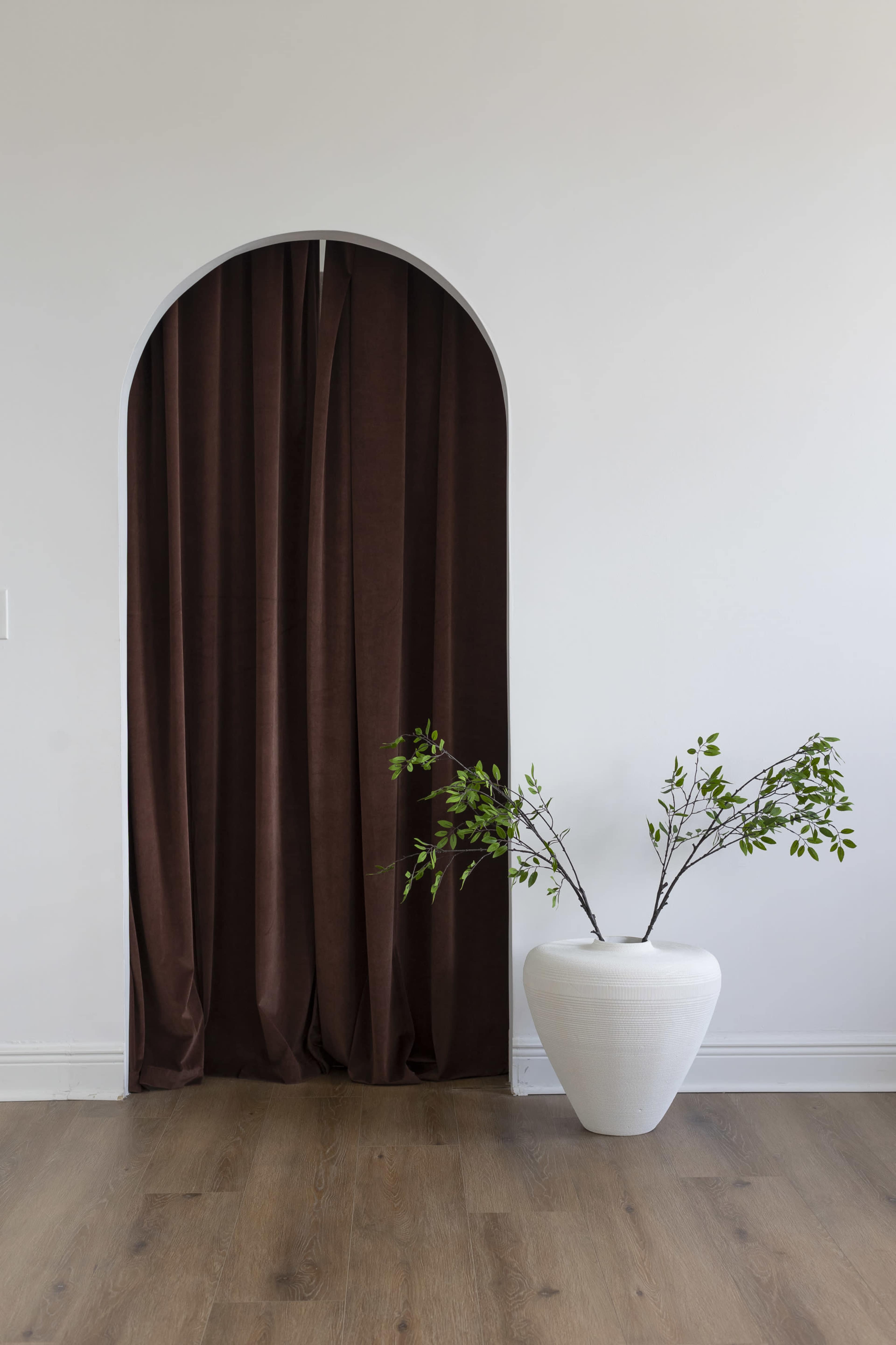 A brown curtain is draped in an arched opening beside a white vase with a green branch against a light wall and wooden floor.