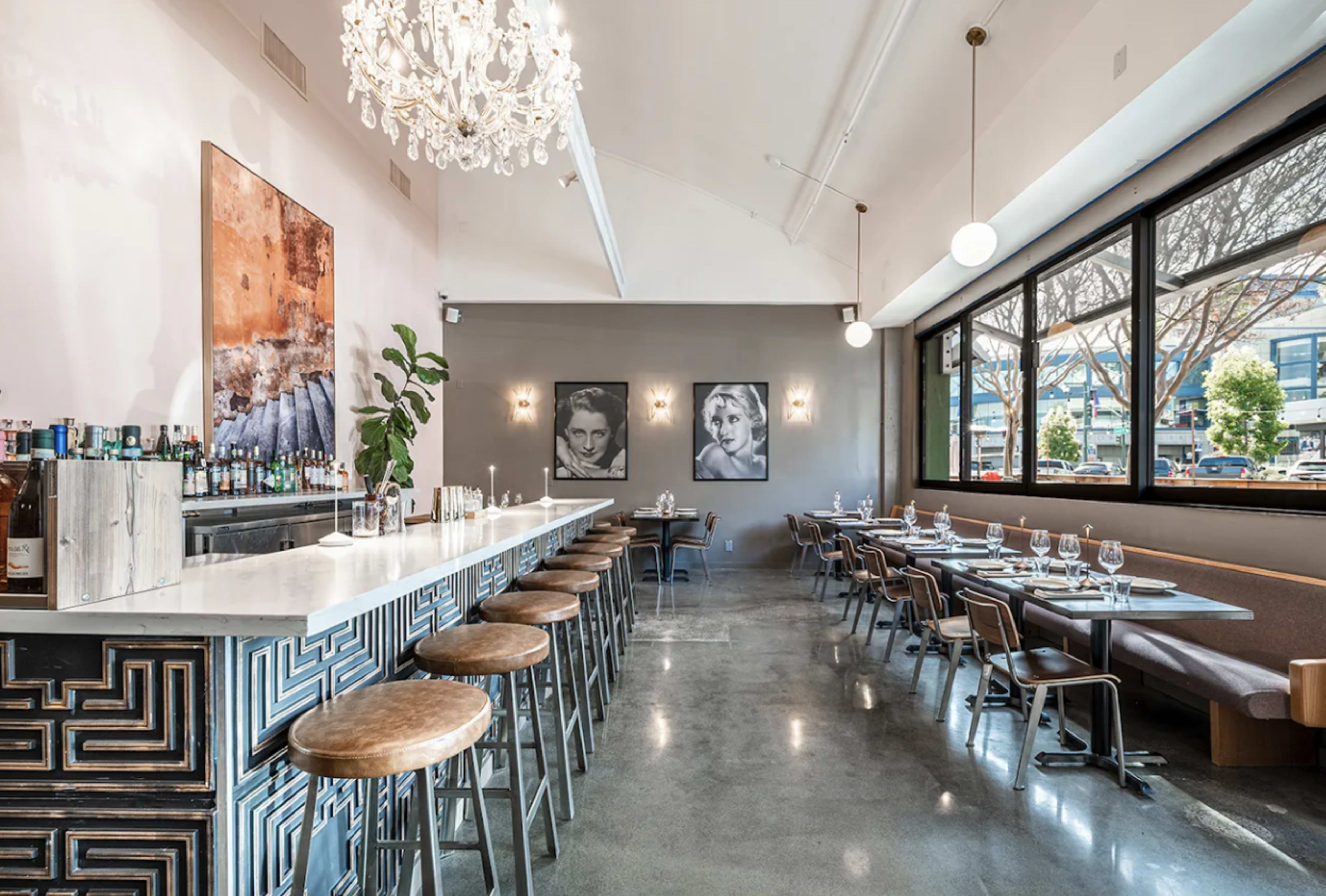 The image shows a modern restaurant interior featuring a bar with high stools, a white marble countertop, and large windows with seating arrangements on either side.