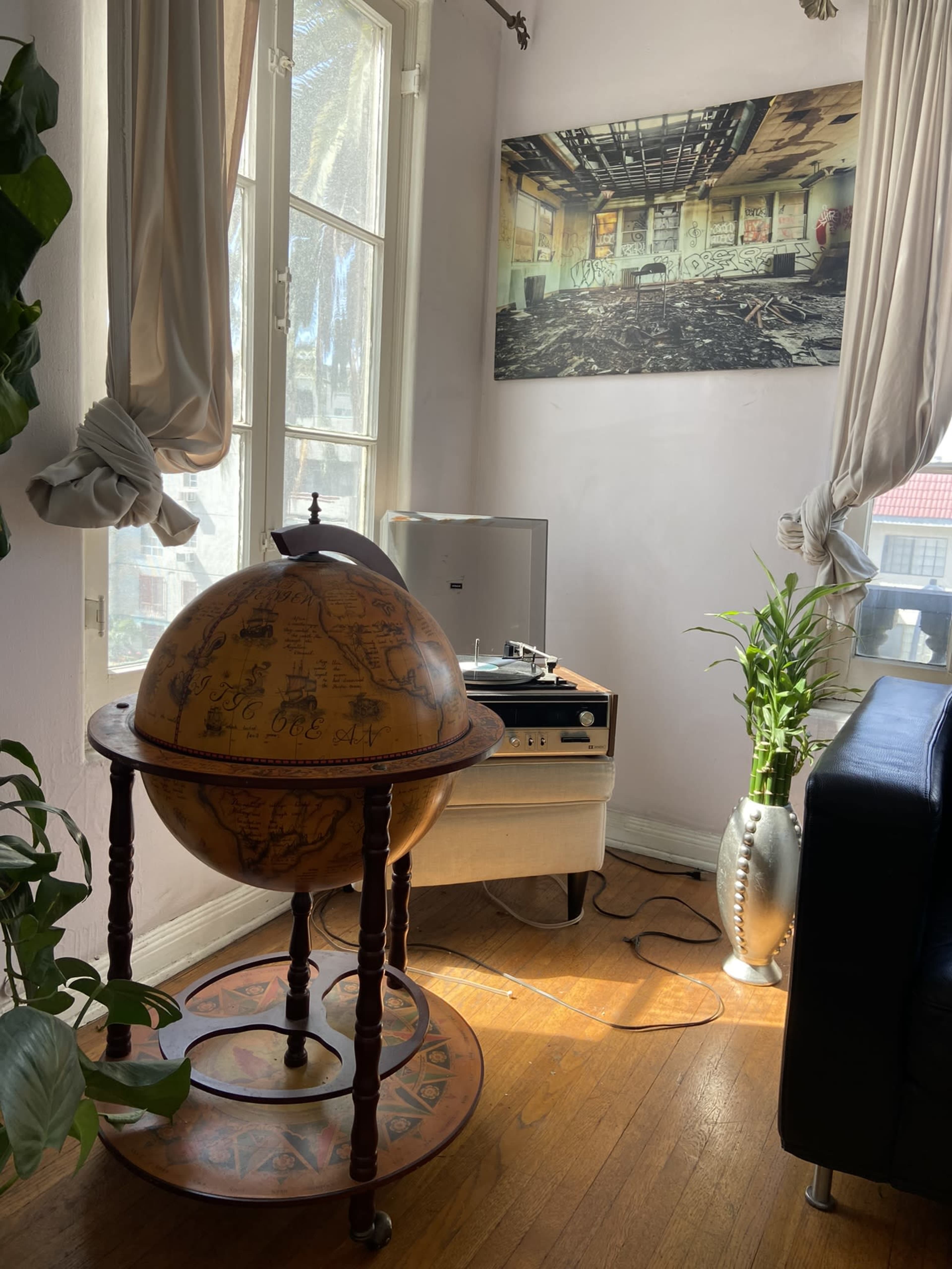 The scene features a vintage globe on a stand, positioned beside a record player and a potted plant in a sunlit corner of a room adorned with floor-to-ceiling windows and a large wall photograph.