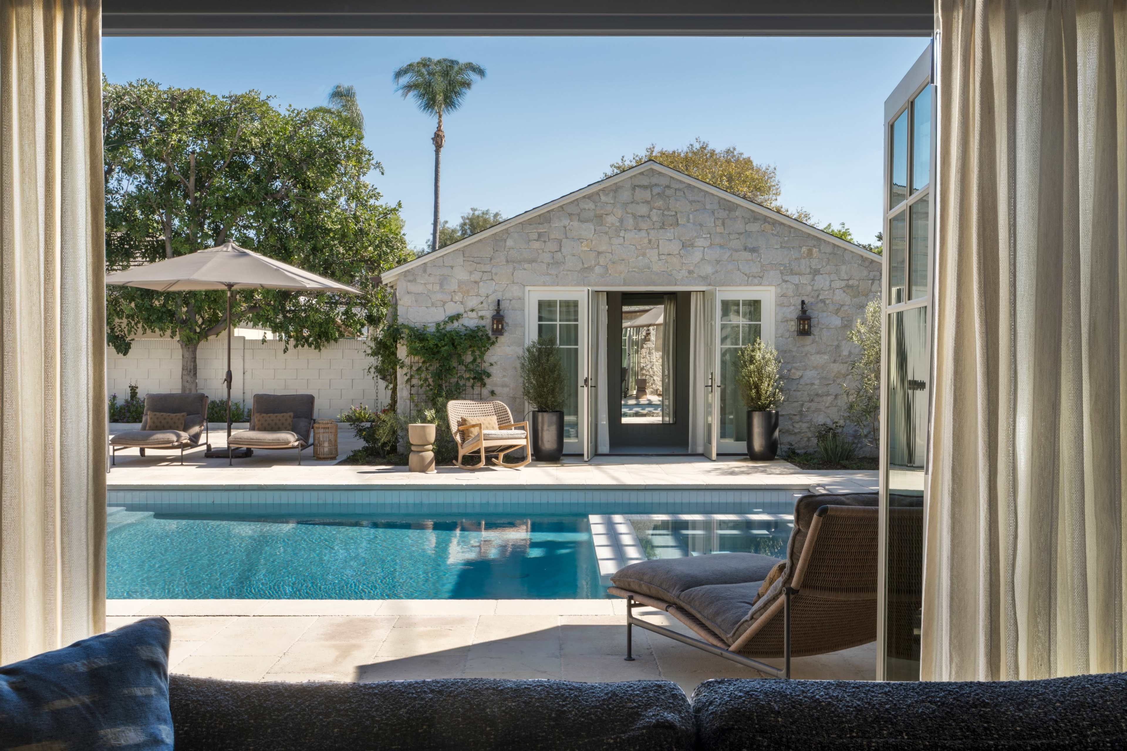 A serene view of a swimming pool surrounded by lounge chairs and a stone house, framed by sheer curtains.