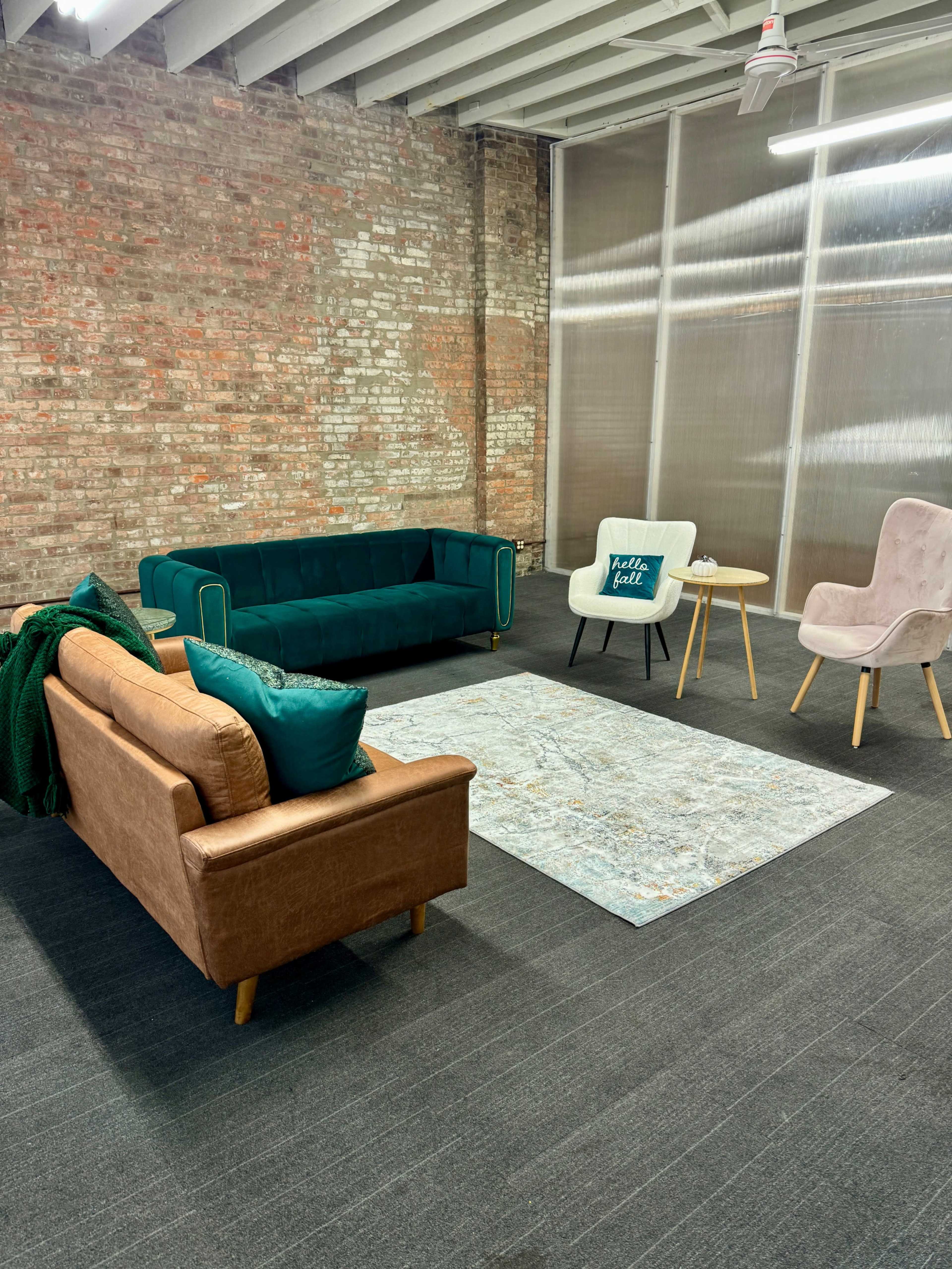 The image shows a modern lounge area featuring a green sofa, a brown couch, two armchairs, and a light rug on a dark carpeted floor, set against a brick wall.