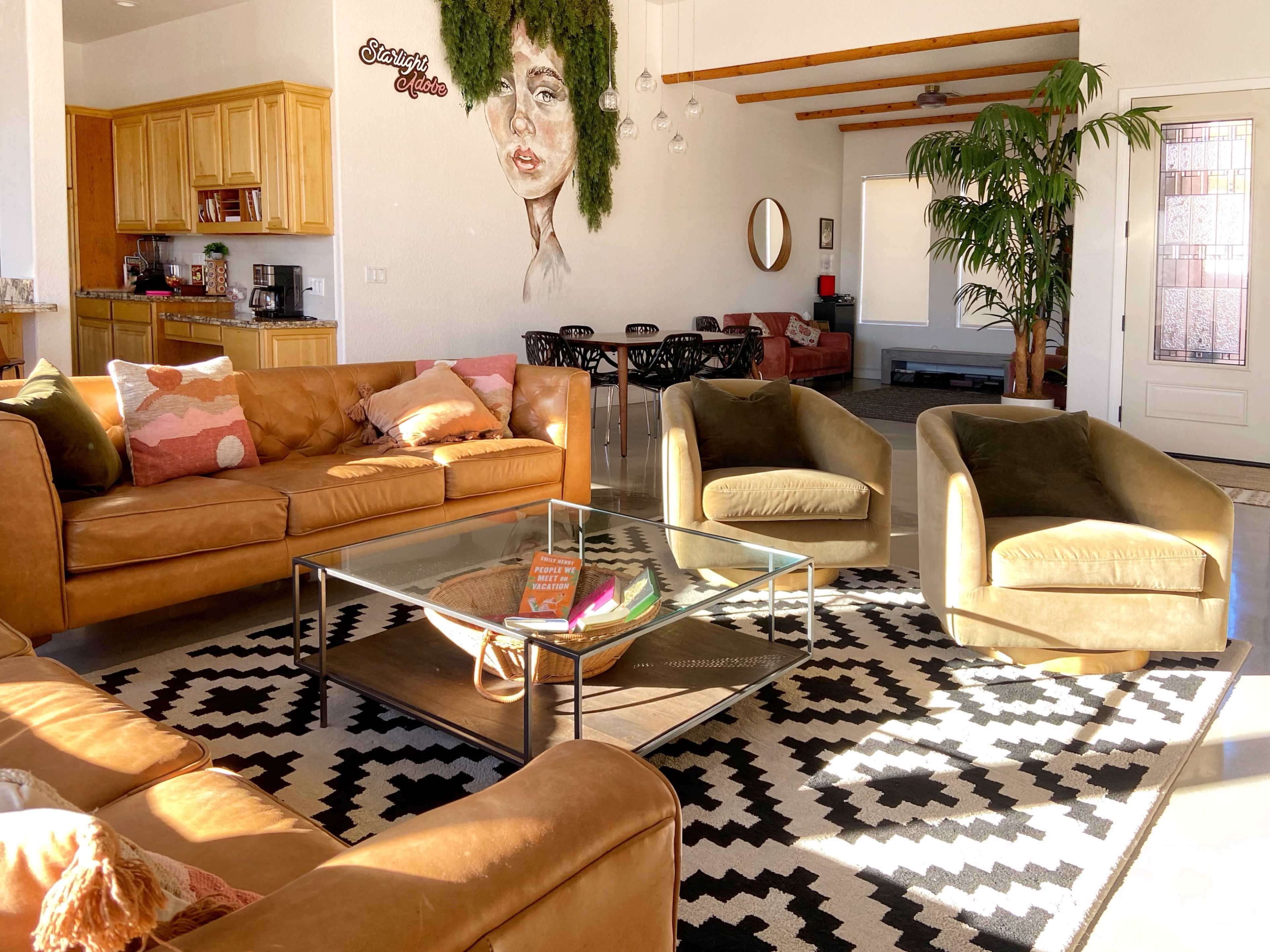 A cozy living room with a brown leather sofa, two armchairs, a glass coffee table, and a large wall mural beside a dining area decorated with plants and modern furniture.