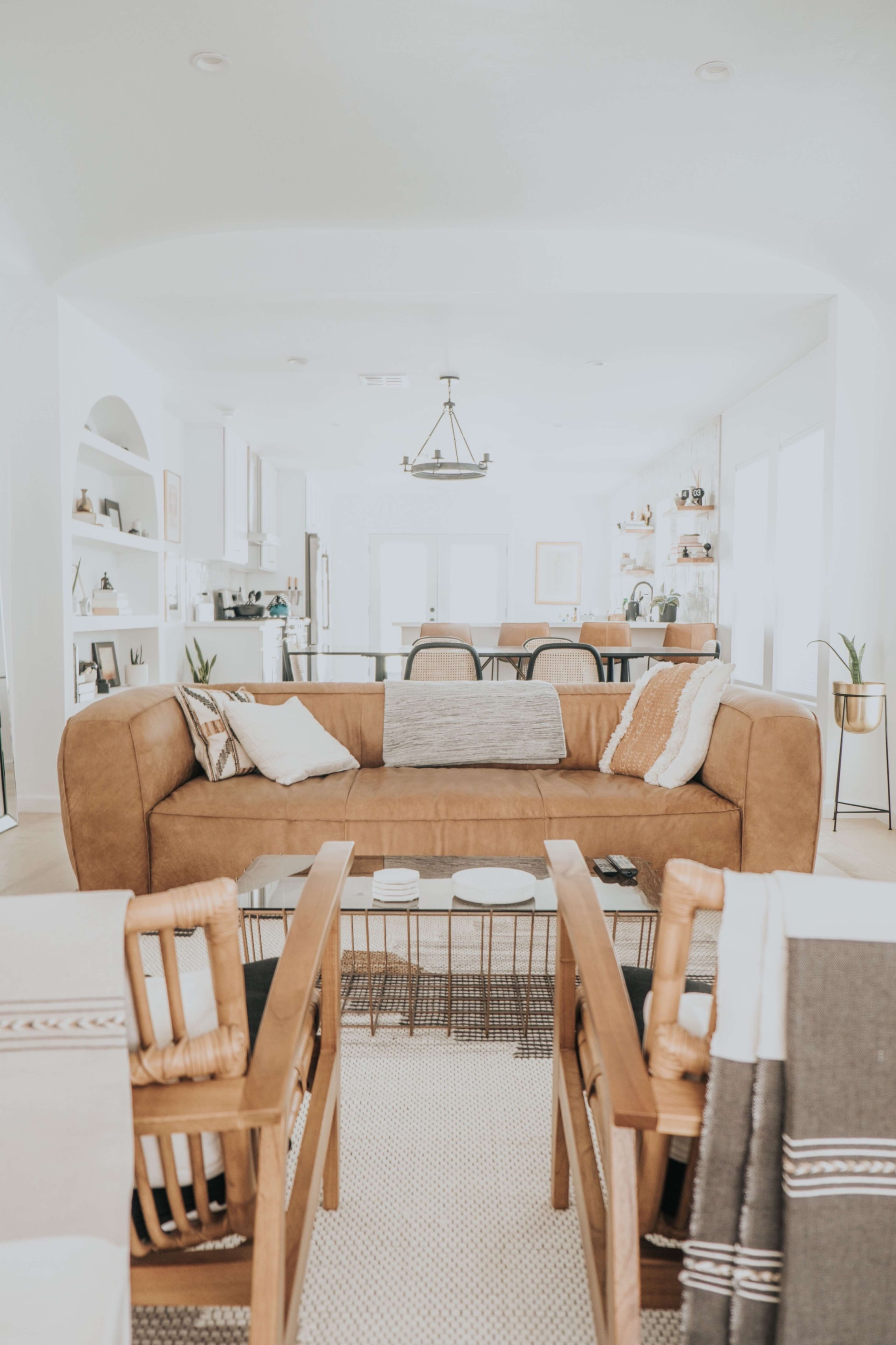 A modern living room features a large brown leather sofa, a coffee table with decorative items, and two wicker chairs, all arranged around a light-colored rug.
