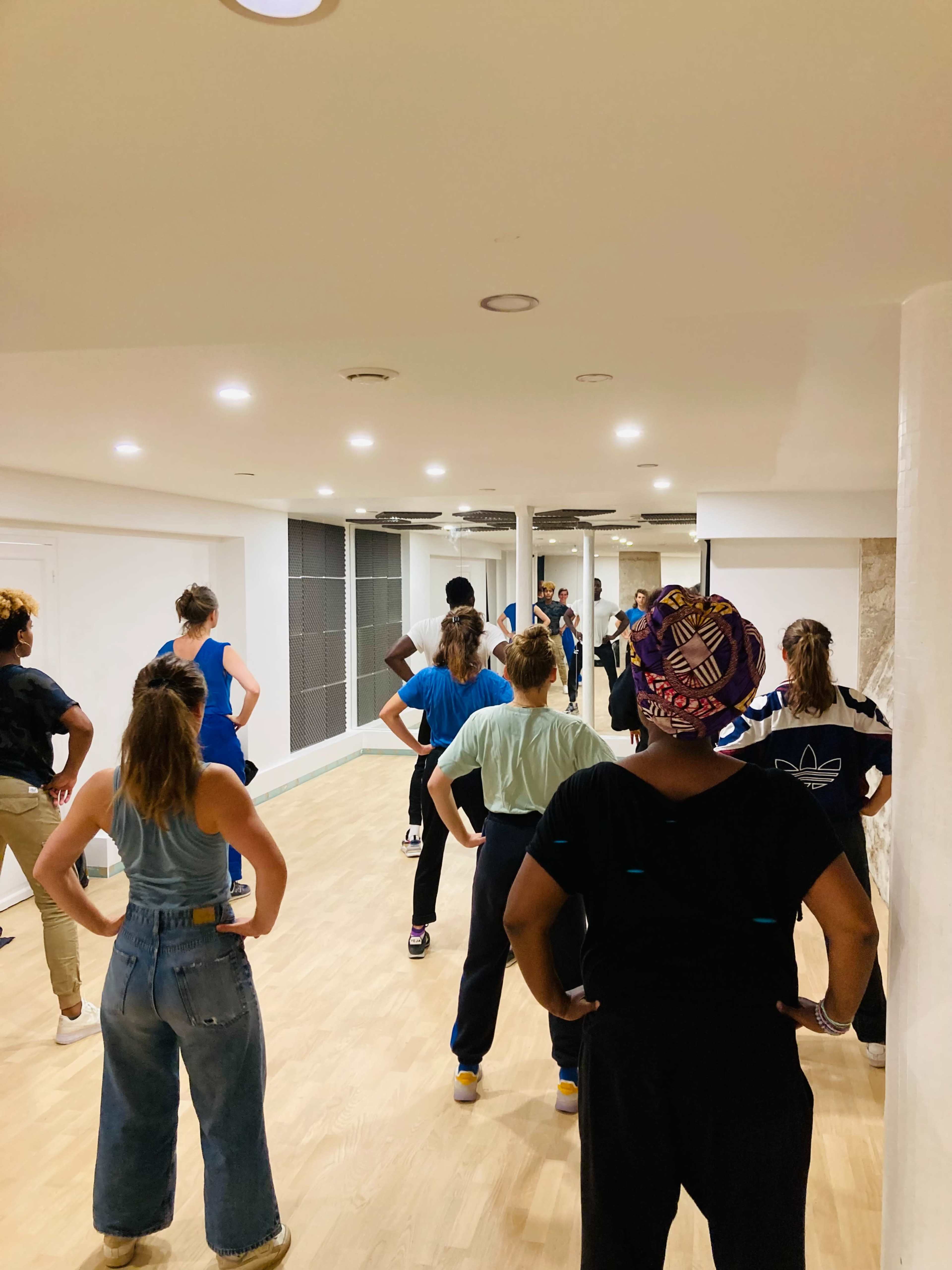 A group of people is practicing dance in a studio with wooden flooring and bright overhead lighting.