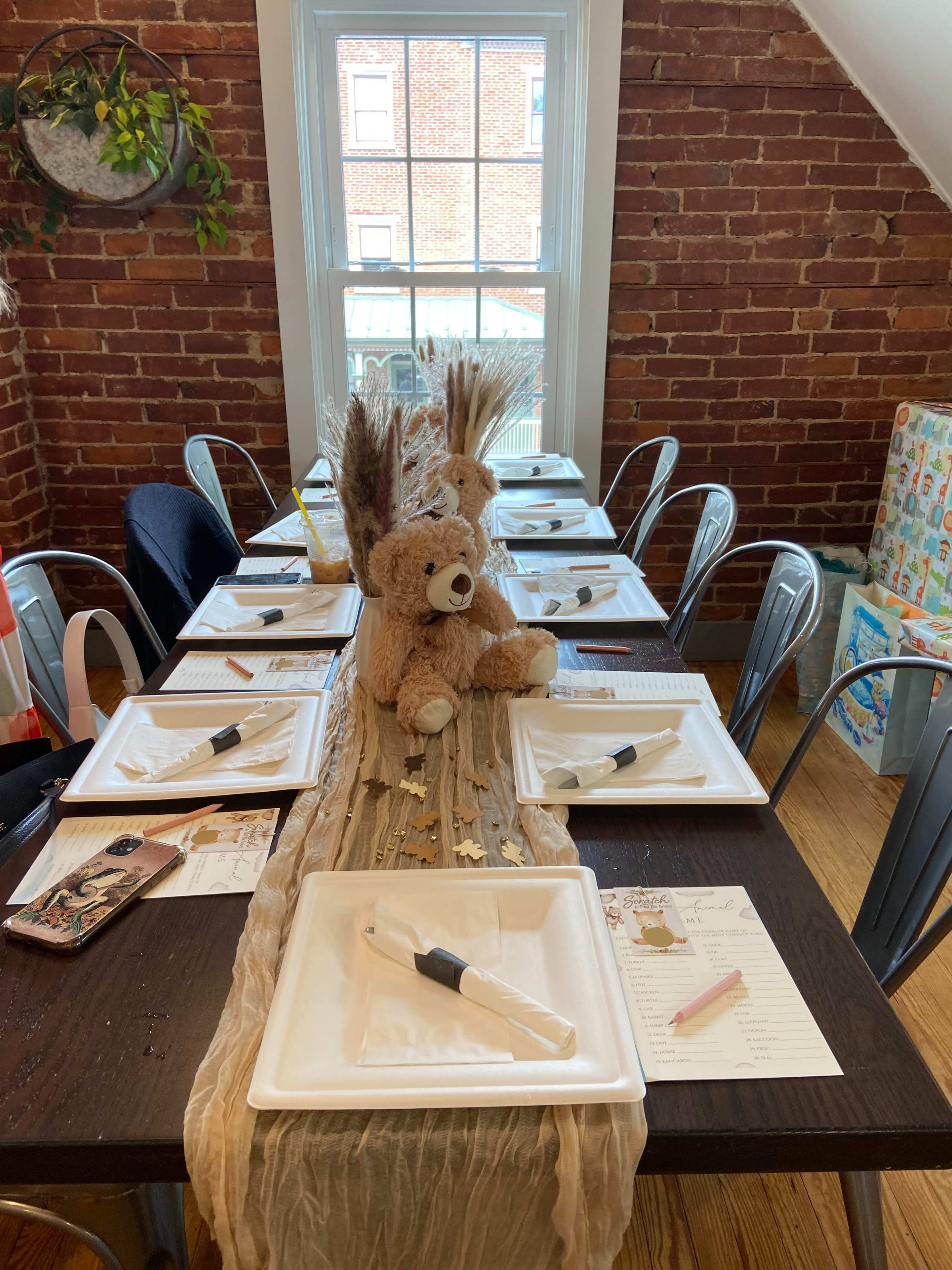 A long table is set with white plates and silverware, featuring a centerpiece of stuffed bears and decorative elements against a backdrop of brick walls and a window.