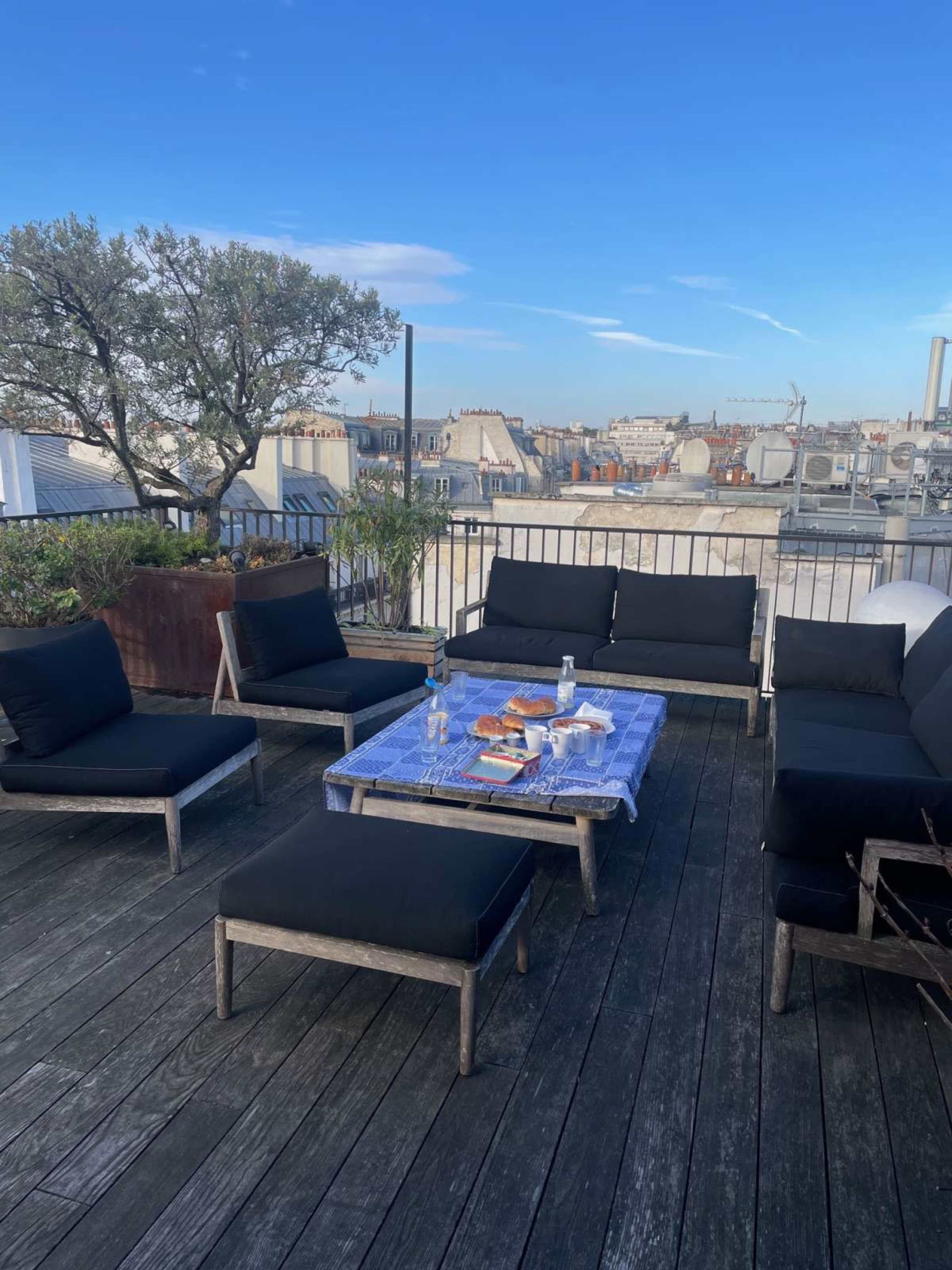 The image shows a rooftop terrace with modern seating arrangements and a coffee table, overlooking a cityscape under a clear blue sky.