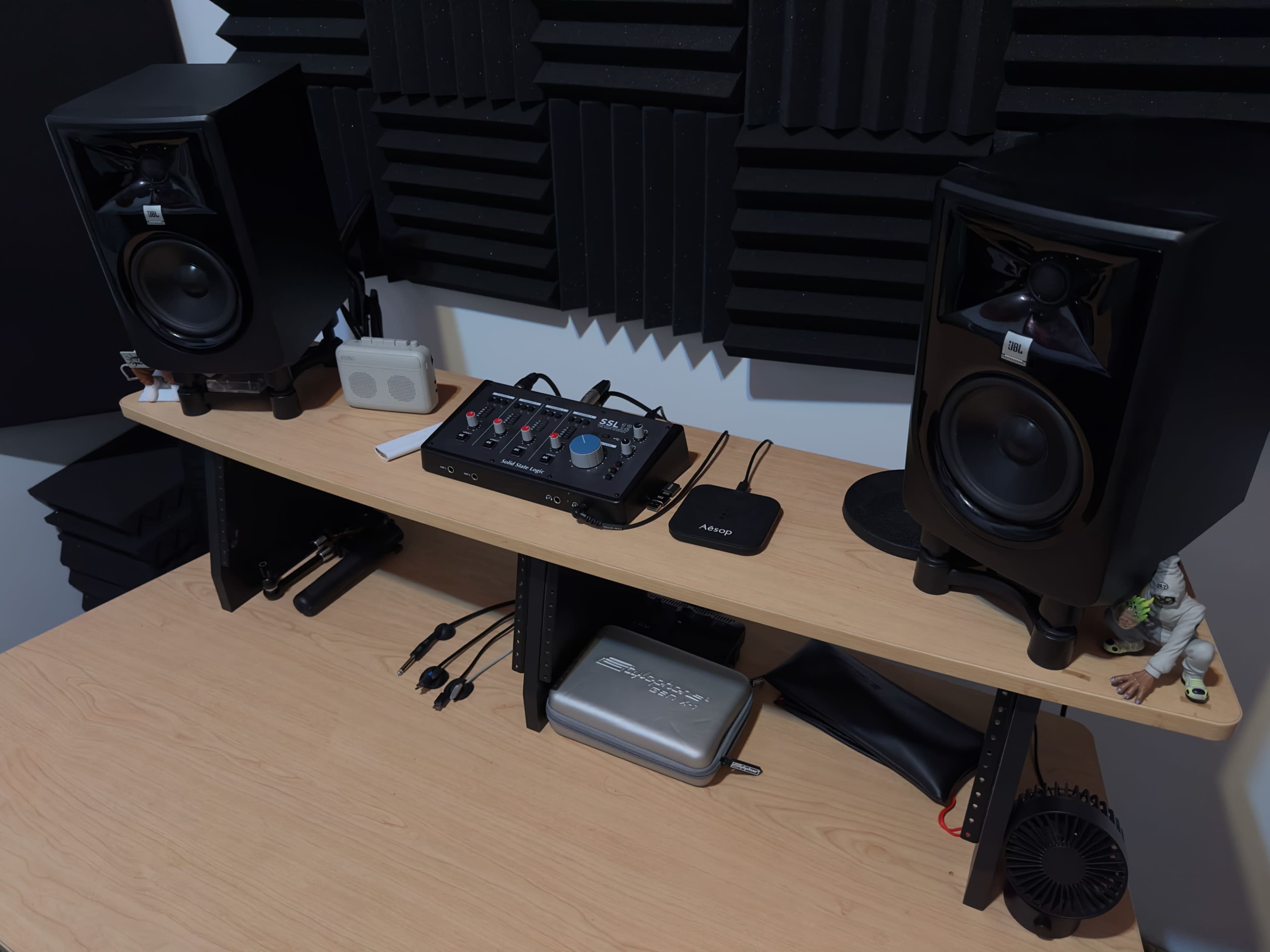 The image shows a sound setup with two black studio monitor speakers, a mixing console, and various audio cables on a wooden desk, surrounded by acoustic foam panels.