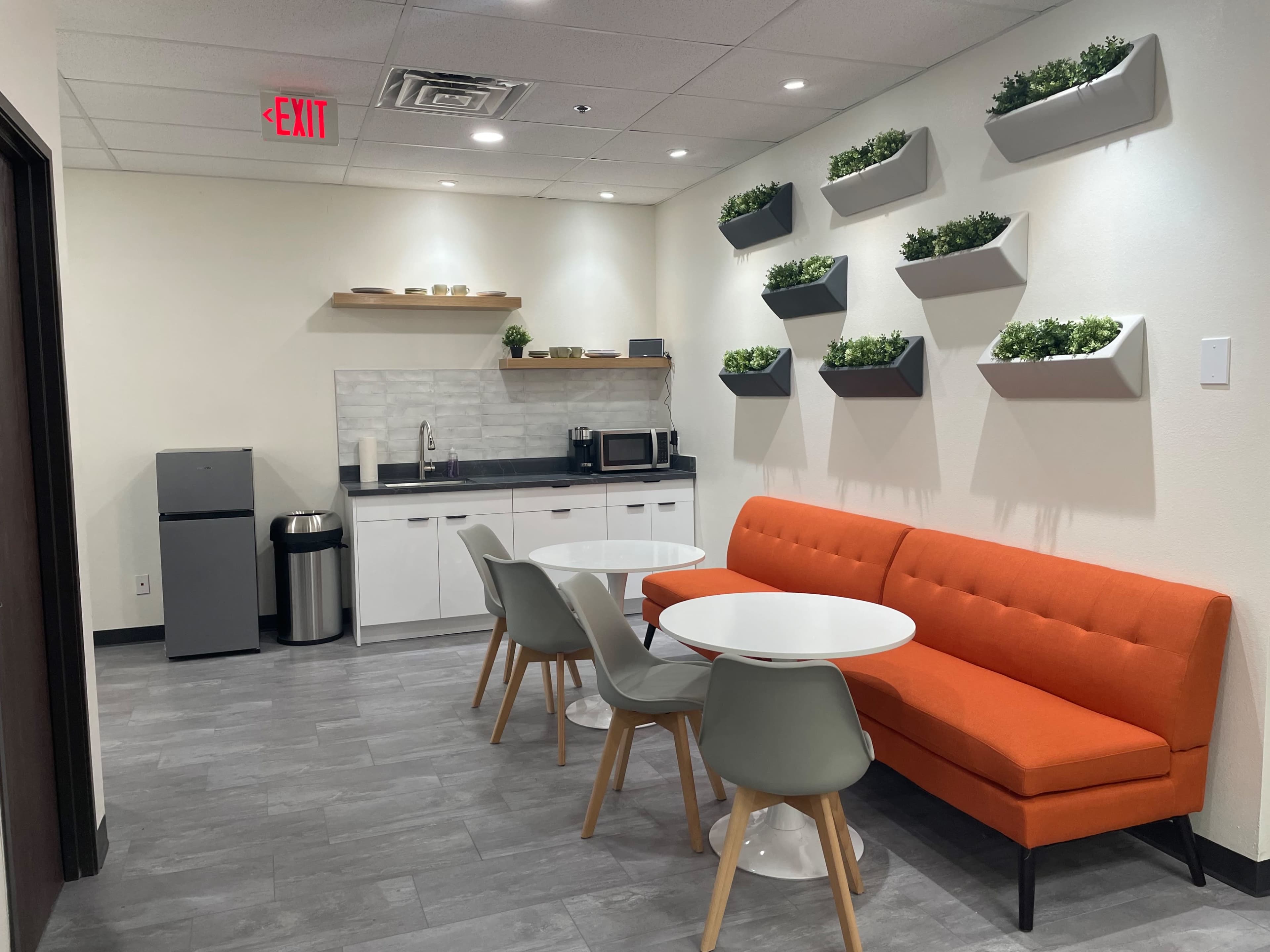 A modern break room featuring an orange couch, white tables, a kitchenette with appliances, and wall-mounted planters.