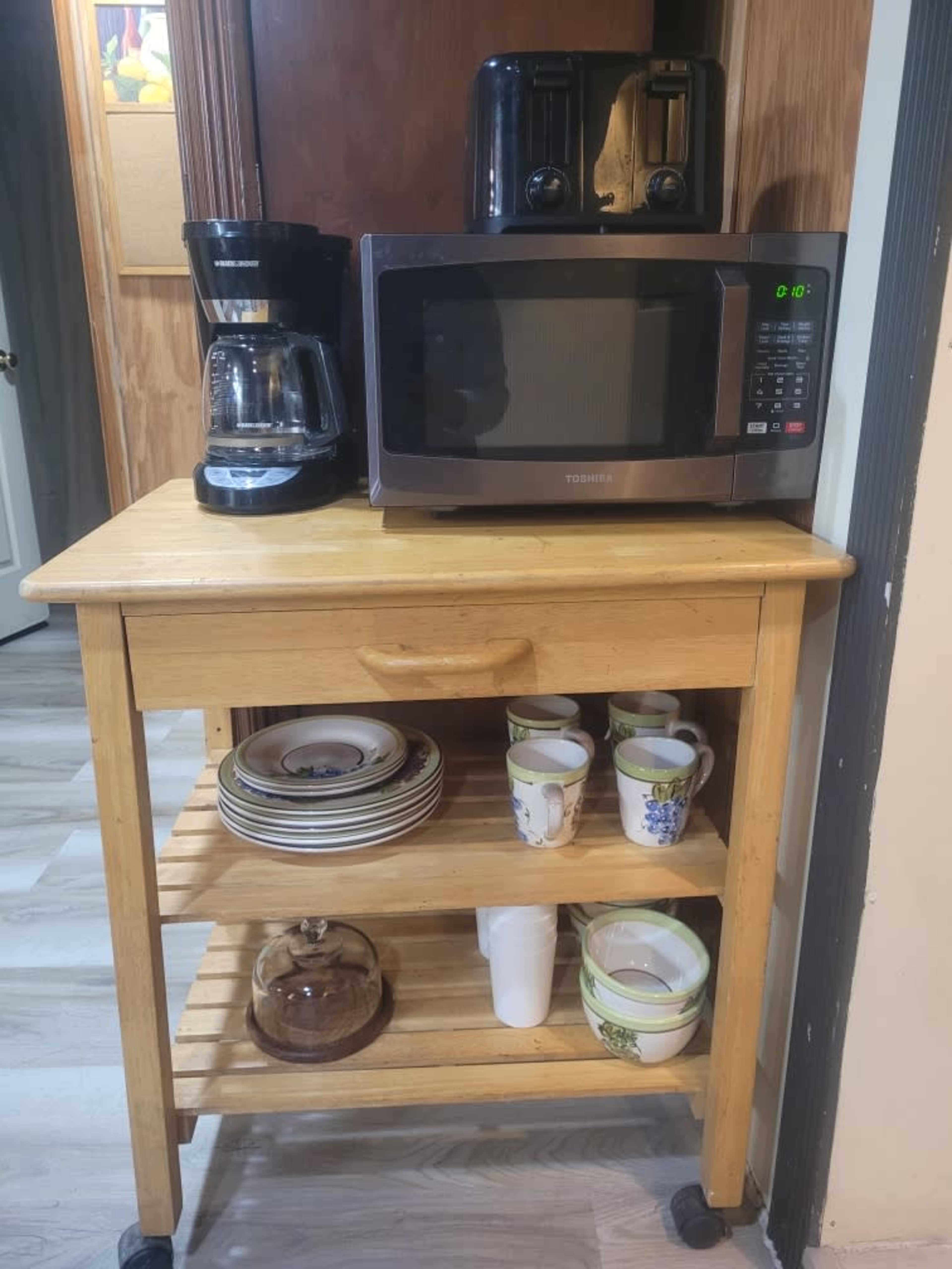 The image shows a wooden kitchen cart with a microwave, coffee maker, and toaster on top, and plates and cups on the shelves below.