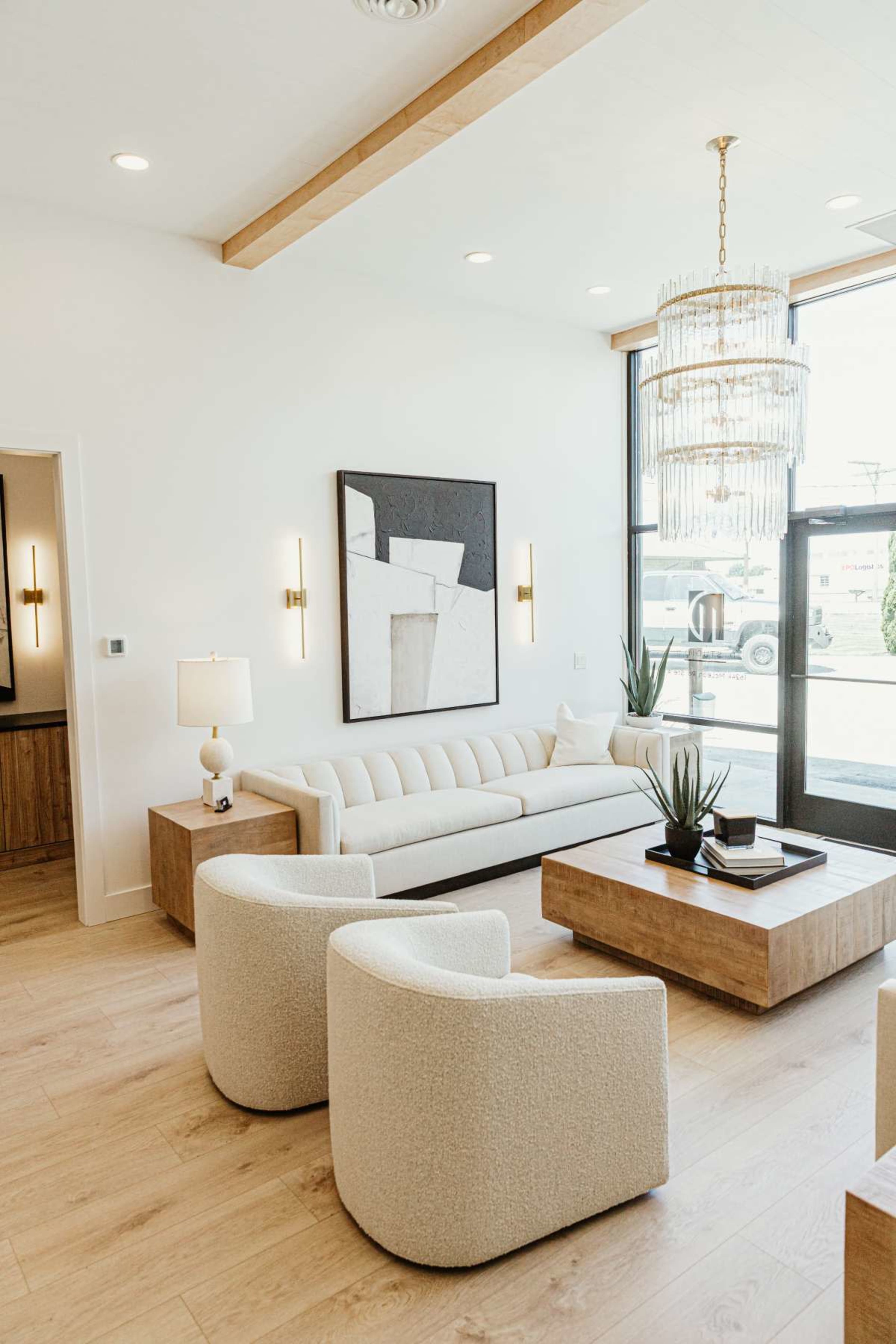 The image shows a modern living room featuring a beige sofa, two round armchairs, a wooden coffee table, and a chandelier, with large windows allowing natural light in.