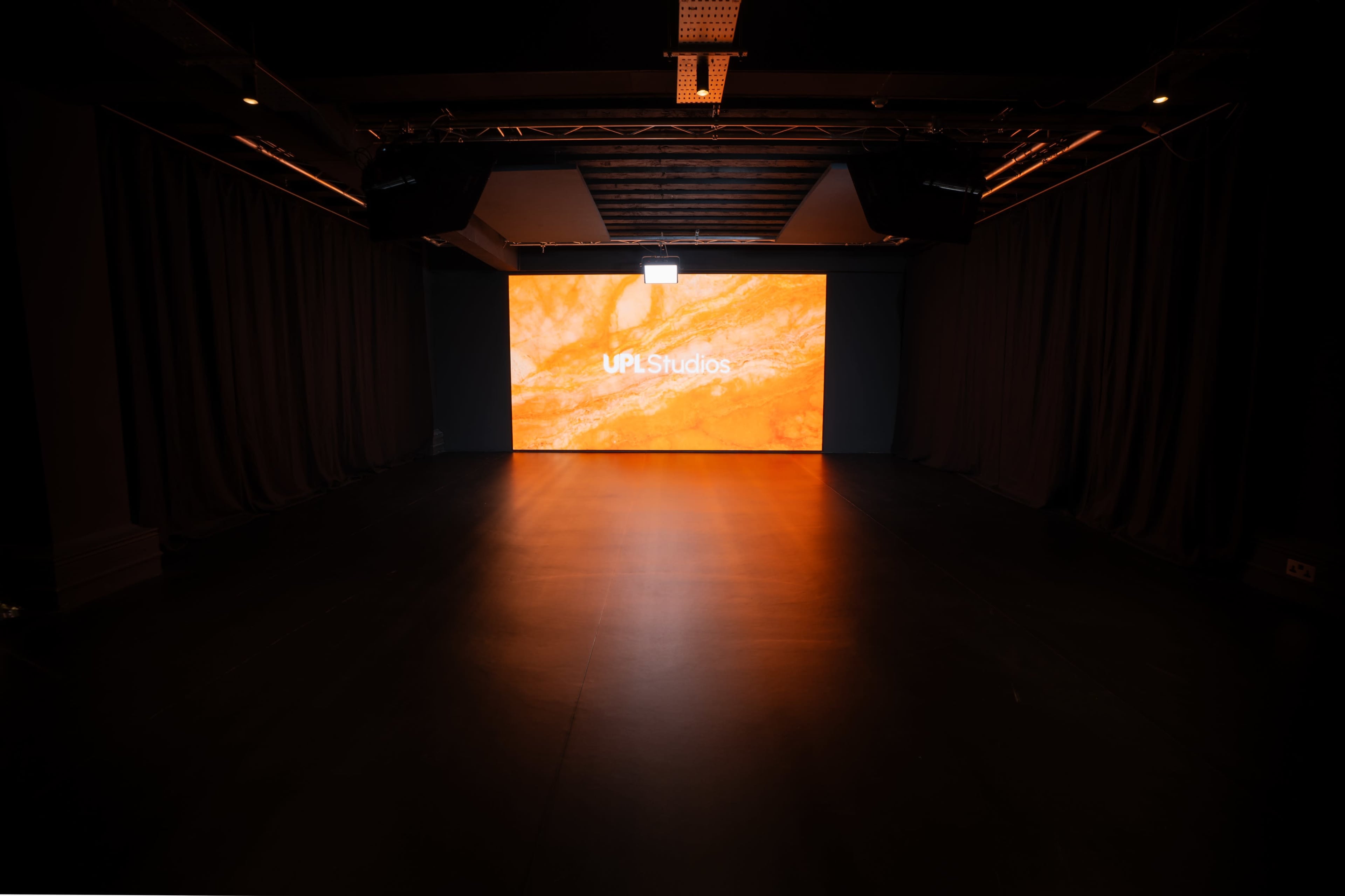 A large event space features a central screen displaying the text "UPLstudios" against a vibrant orange background, with dark curtains lining the sides.