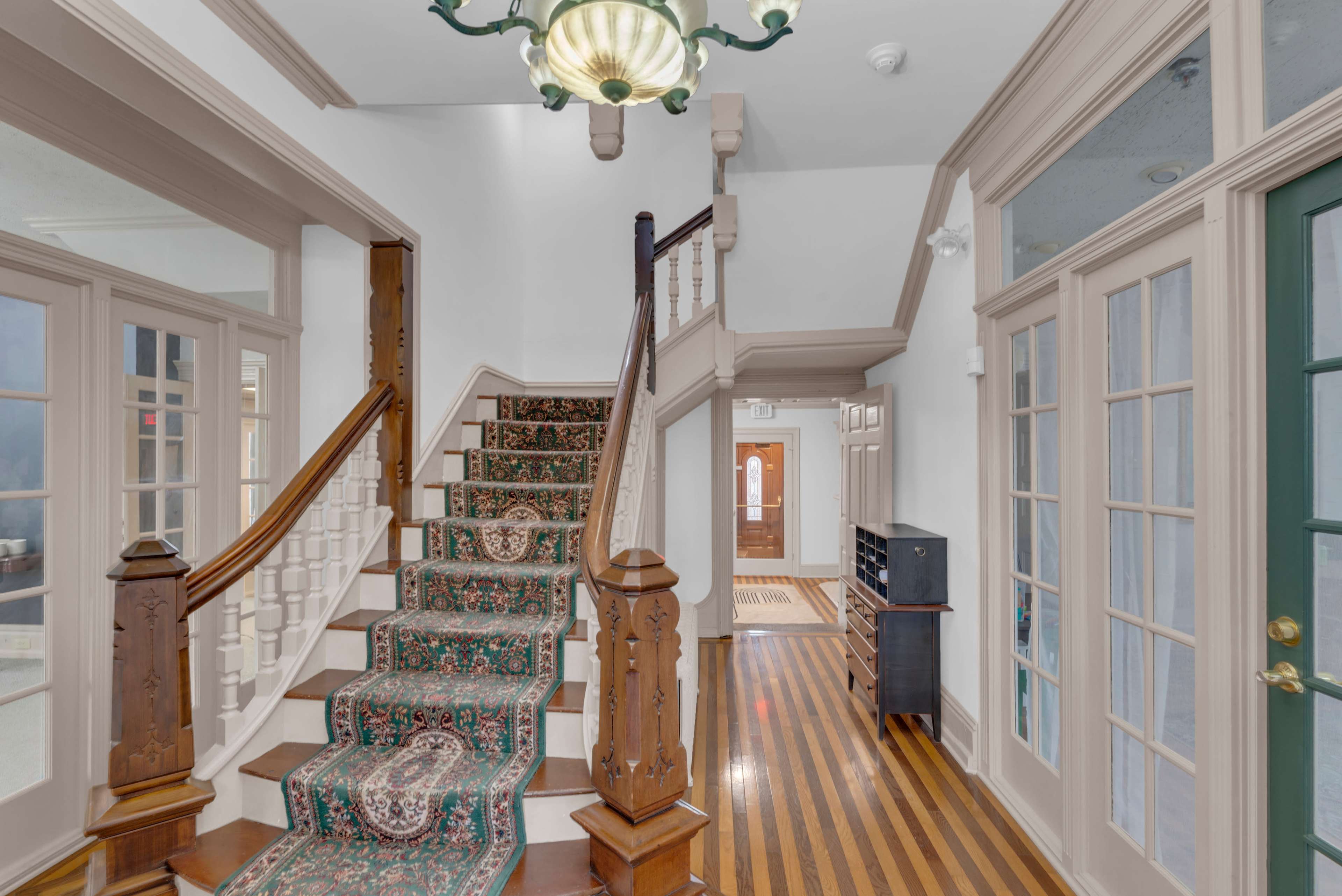 The image shows a well-lit foyer featuring a staircase with a patterned carpet, wooden railings, and a chandelier overhead, alongside multiple glass-paneled doors.