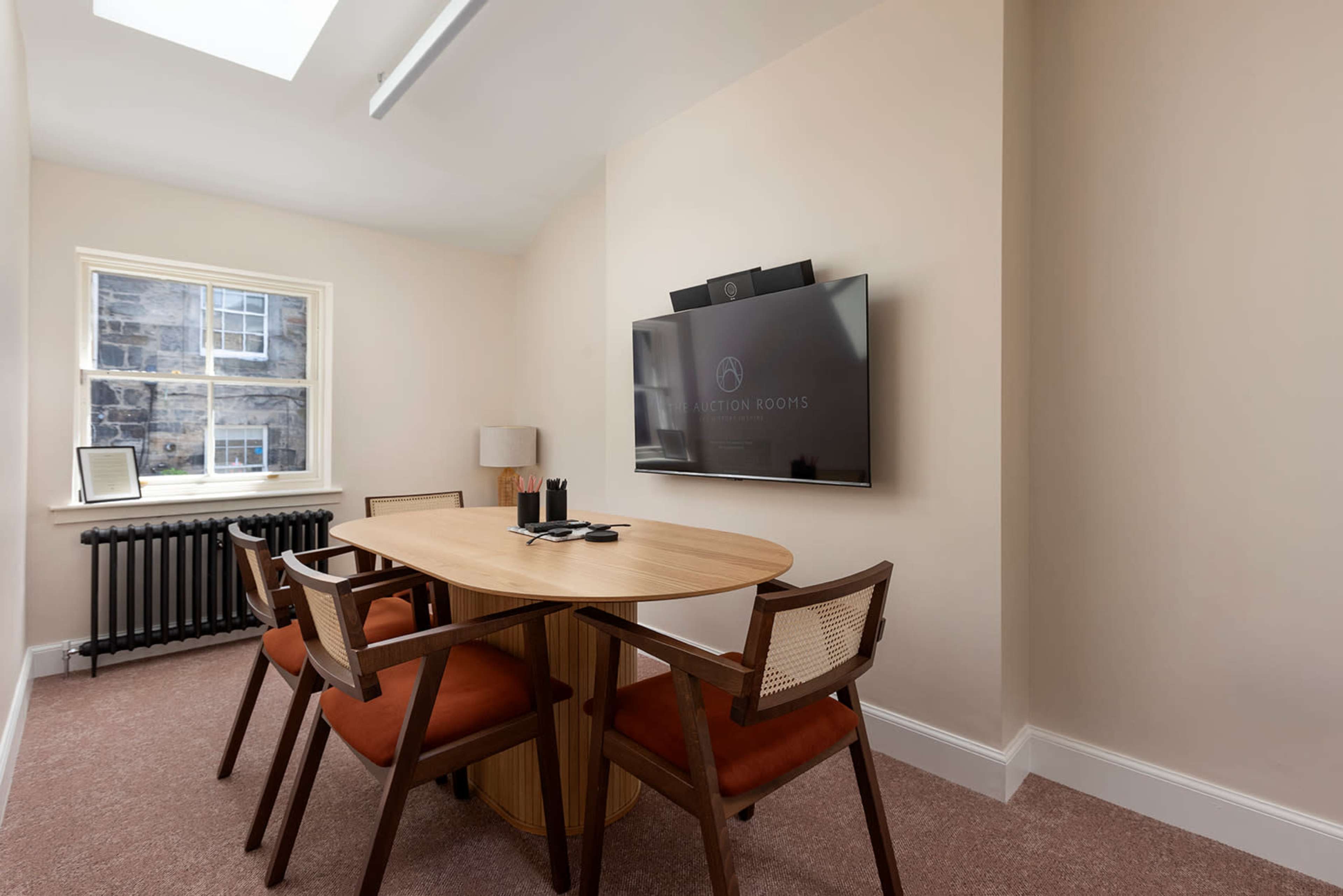 A small meeting room features a round wooden table with four chairs, a wall-mounted television, and a window allowing natural light to enter.