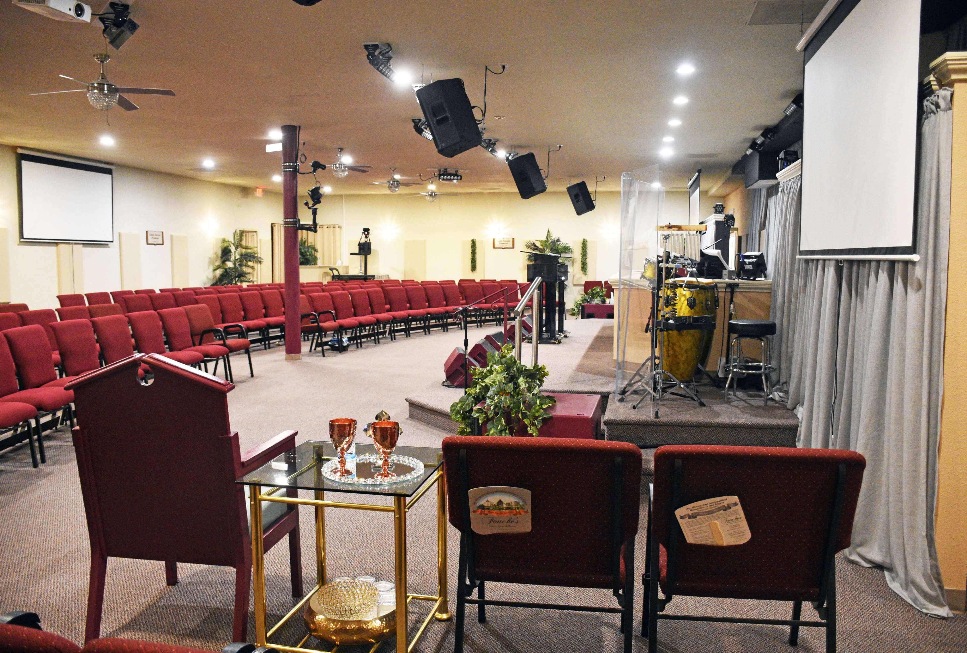 A nearly empty church auditorium with rows of red upholstered chairs, a stage featuring musical instruments, and a small table set with decorative items in the foreground.
