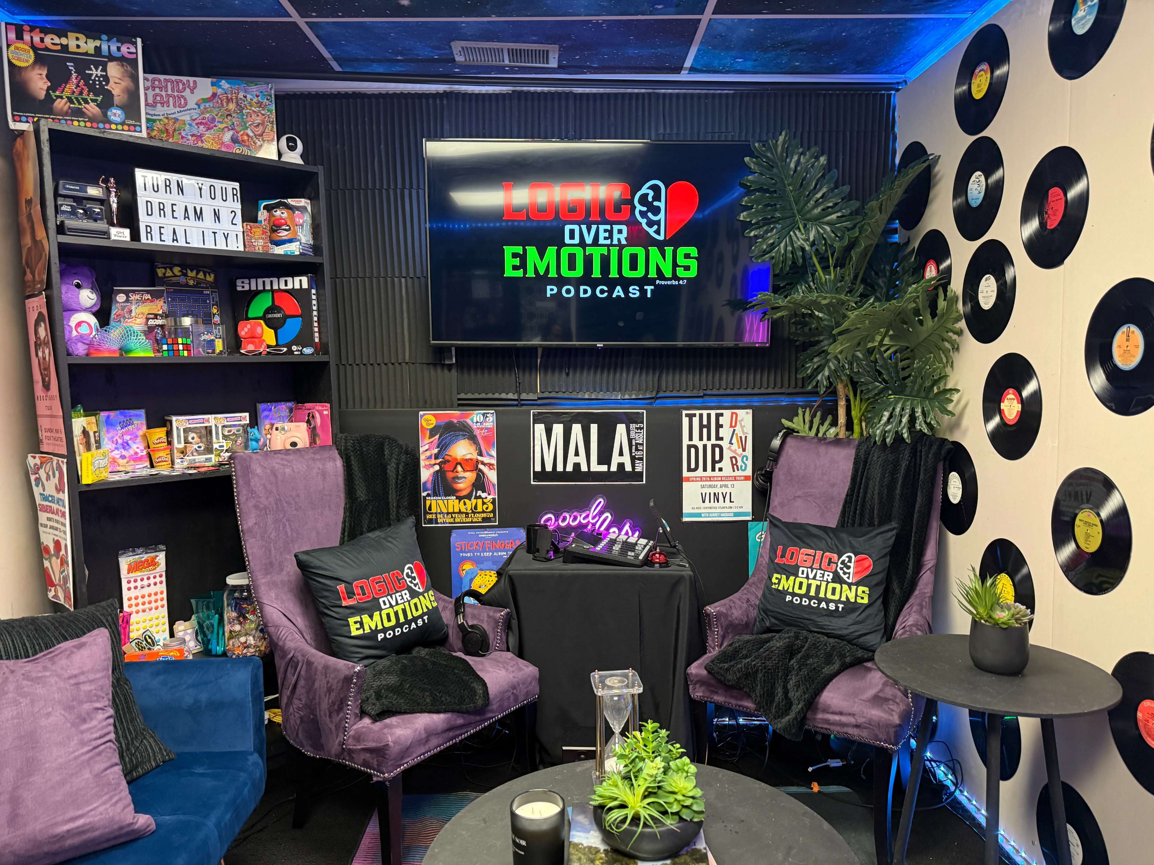The image shows a colorful podcast studio decorated with vinyl records, a television displaying the podcast title "Logic Over Emotions," and two purple chairs facing each other beside small tables.