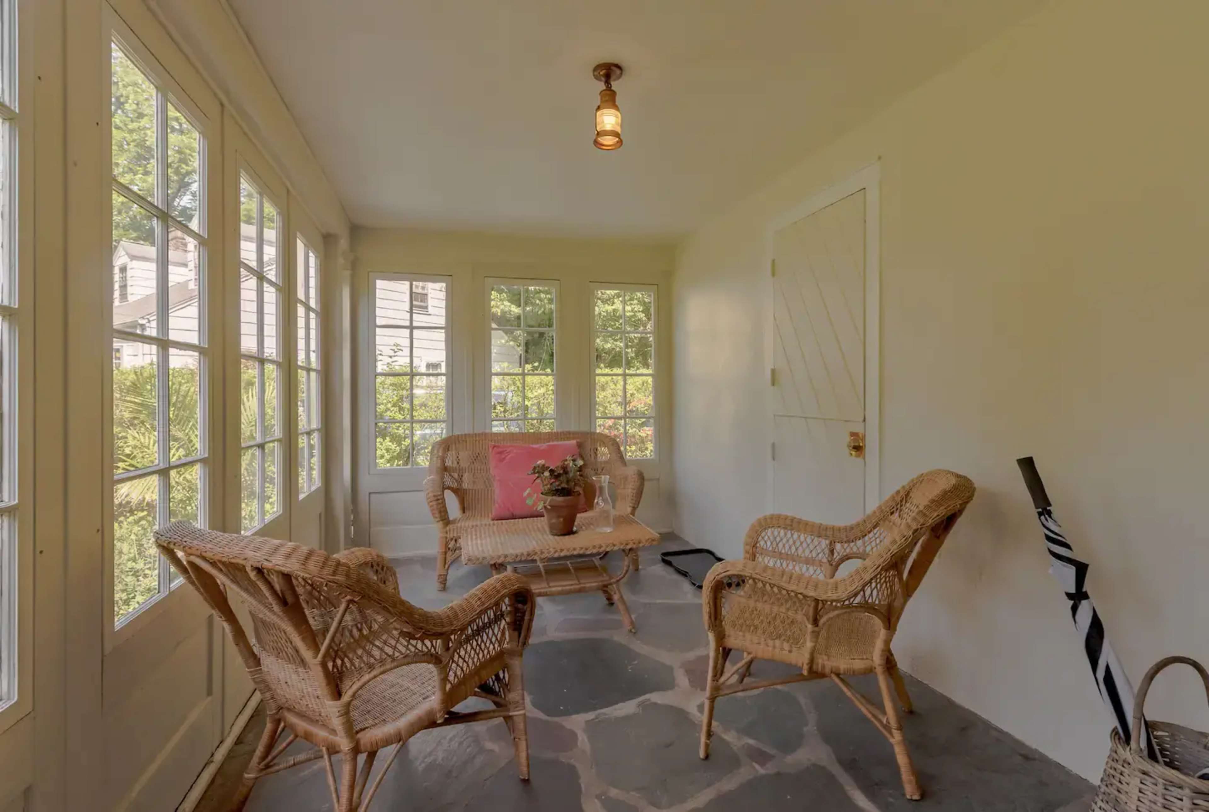 A sunlit enclosed porch featuring a small seating area with wicker furniture and a potted plant.