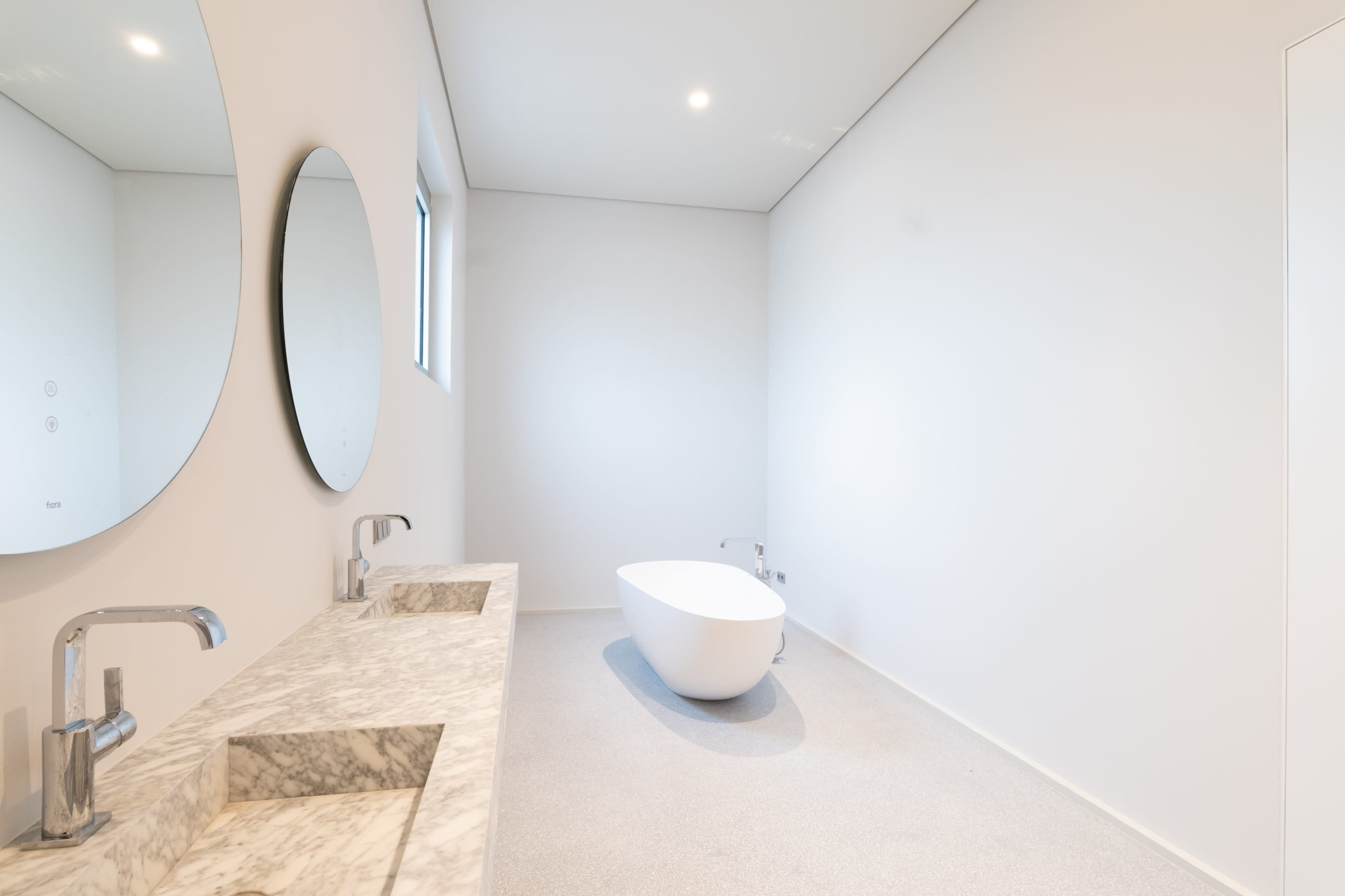 A minimalist bathroom features a freestanding white bathtub and a double sink with a marble countertop and circular mirrors.