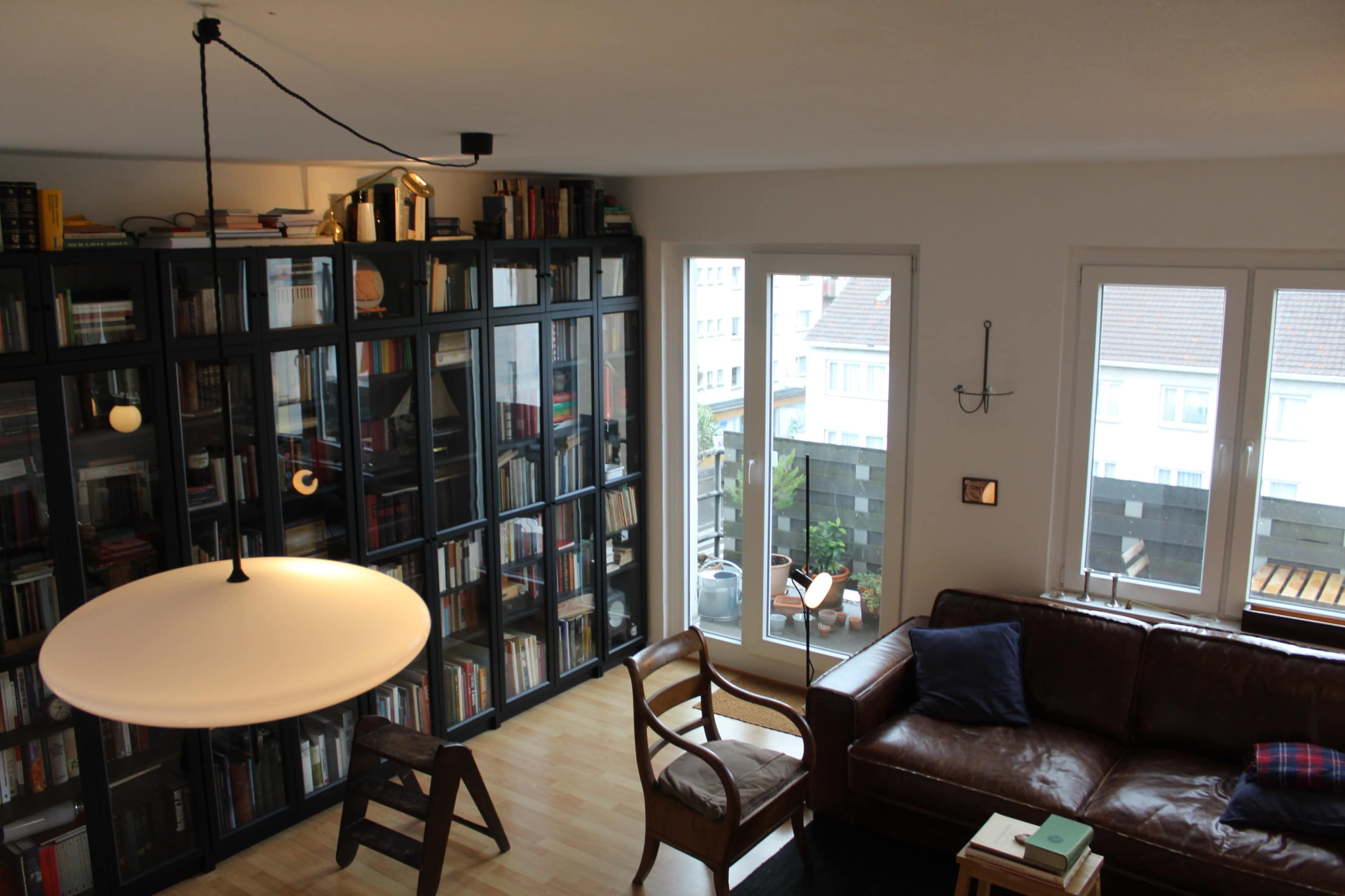 The image shows a living room with a bookshelf filled with books, a brown leather sofa, and large windows overlooking an outdoor area.