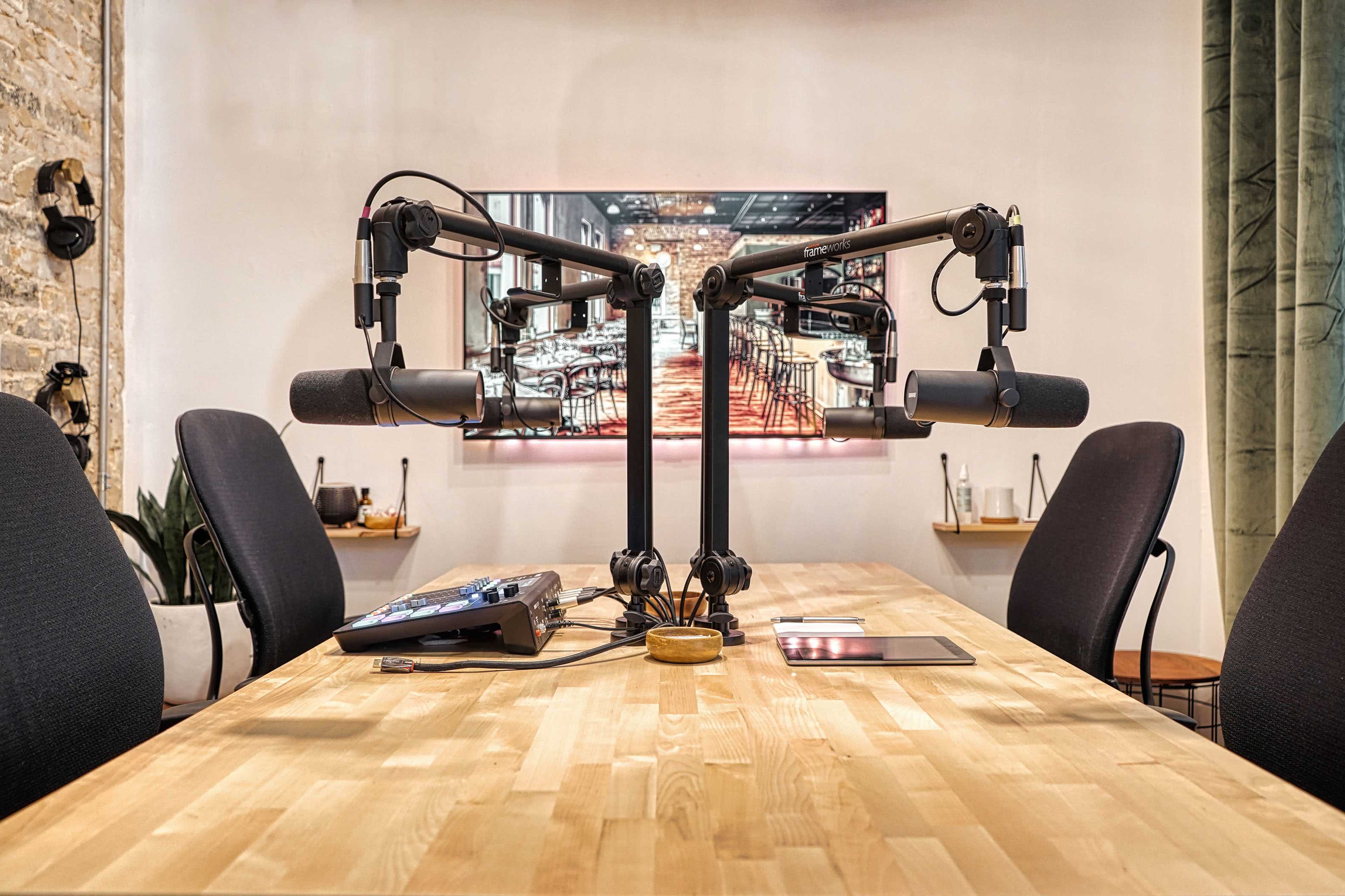 A modern podcast recording studio features two microphones on adjustable arms positioned over a wooden table with a touchscreen device and a circular coaster, set against a backdrop of a dimly lit bar scene on a screen.