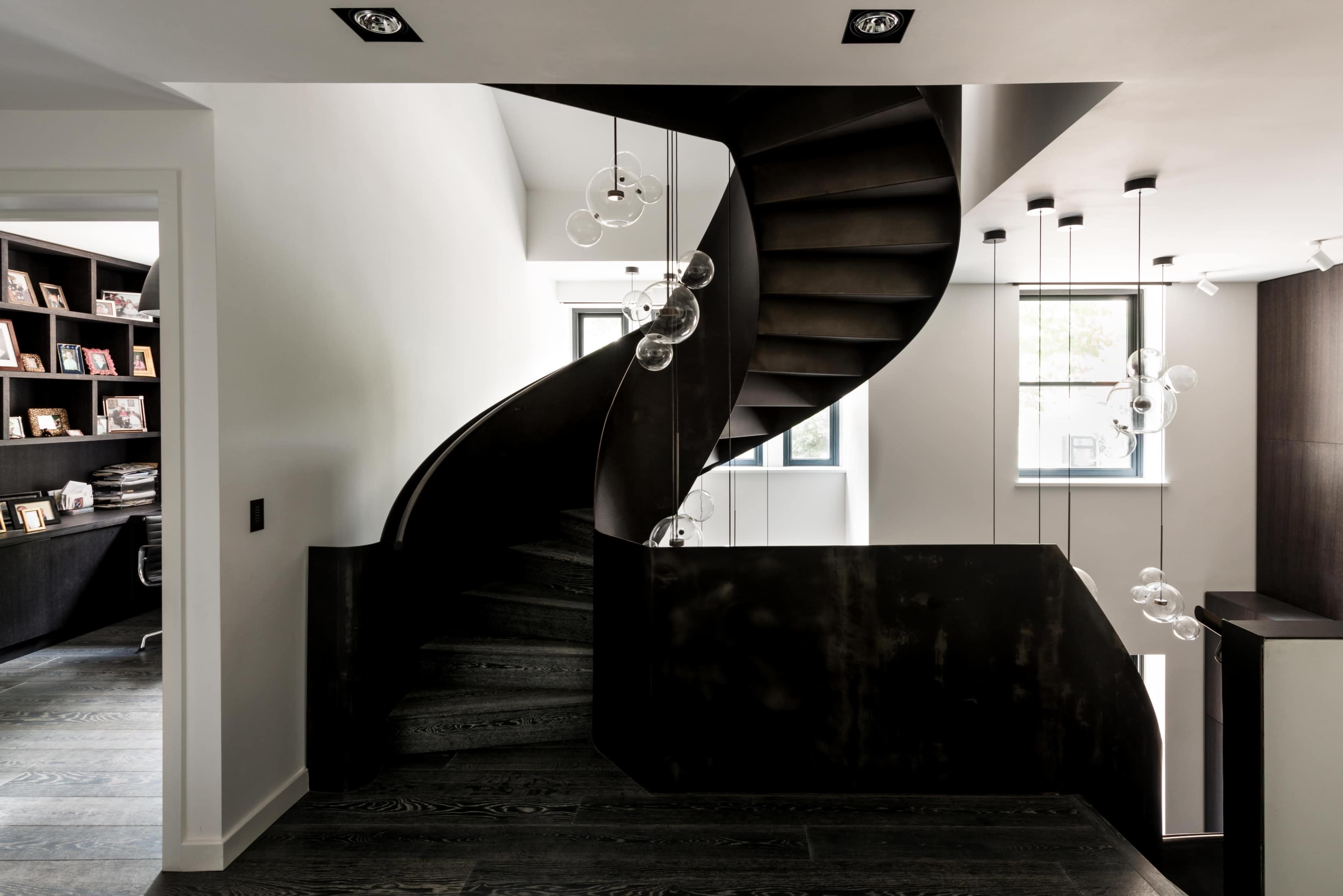 A modern interior features a sweeping black spiral staircase illuminated by pendant lights, with a home office visible in the background.