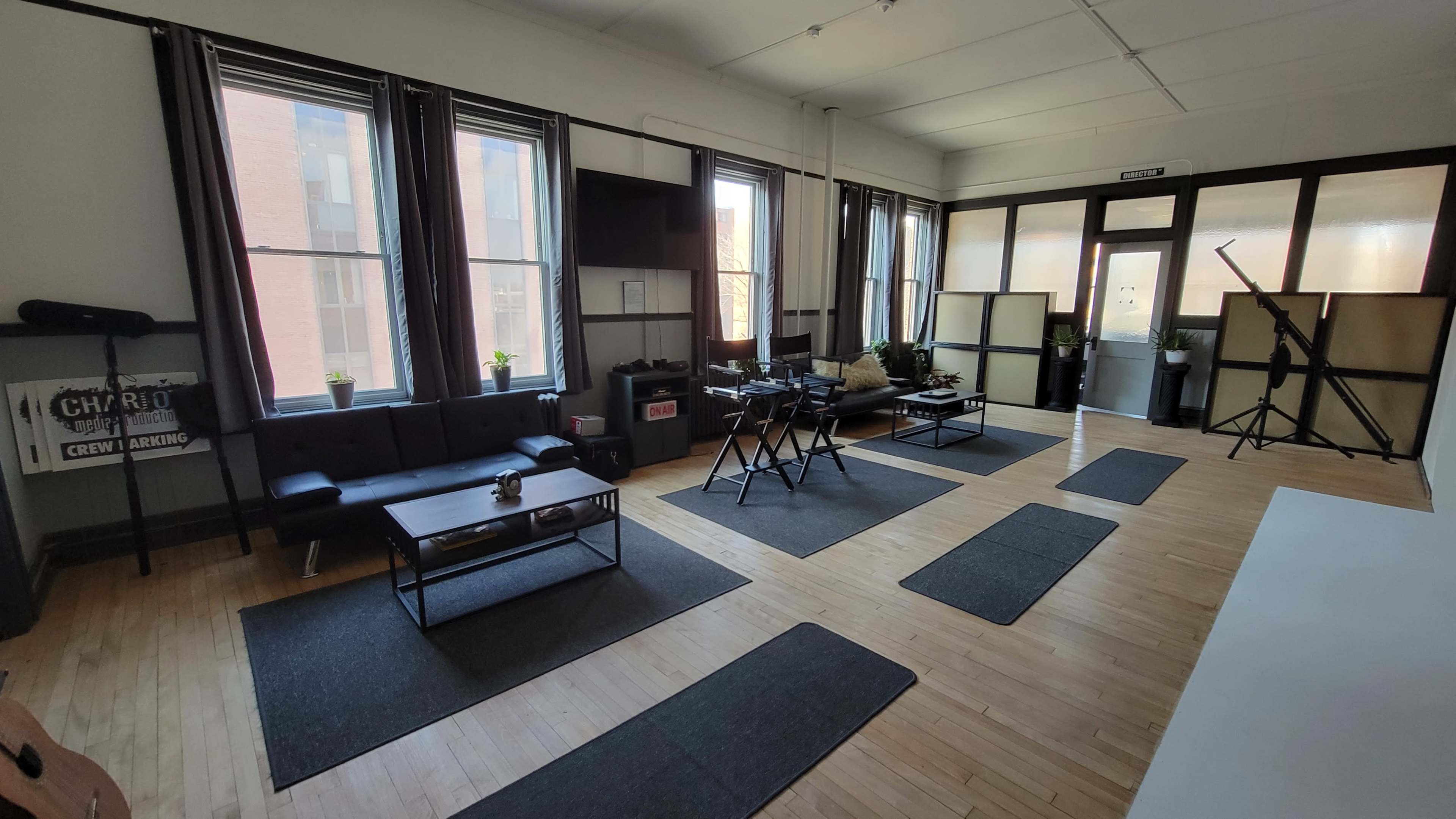 A spacious room with a hardwood floor, featuring large windows, a black couch, a coffee table, and several plants.