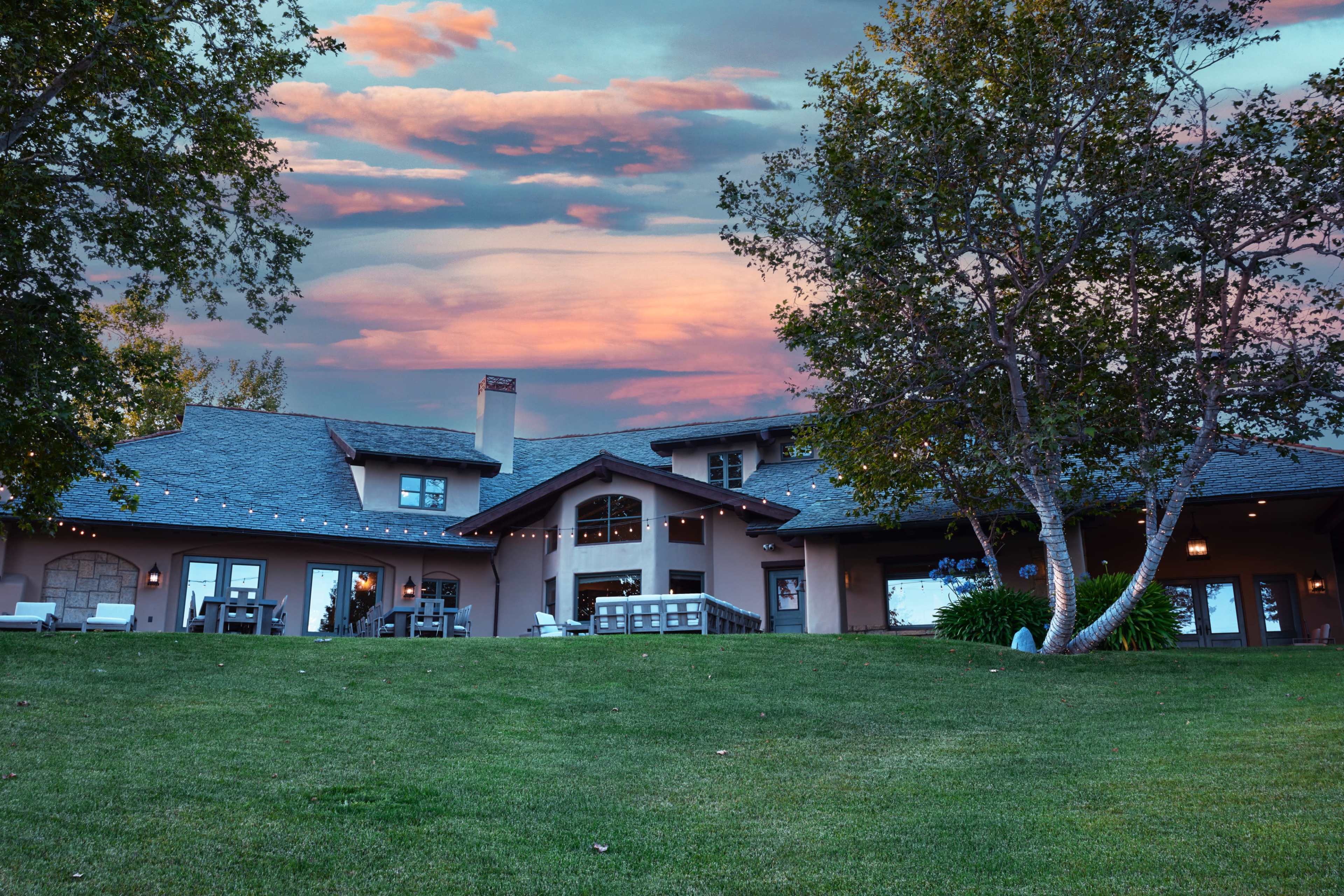 A large, two-story house with a wide porch and multiple windows is set against a colorful sky at sunset, surrounded by green grass and trees.
