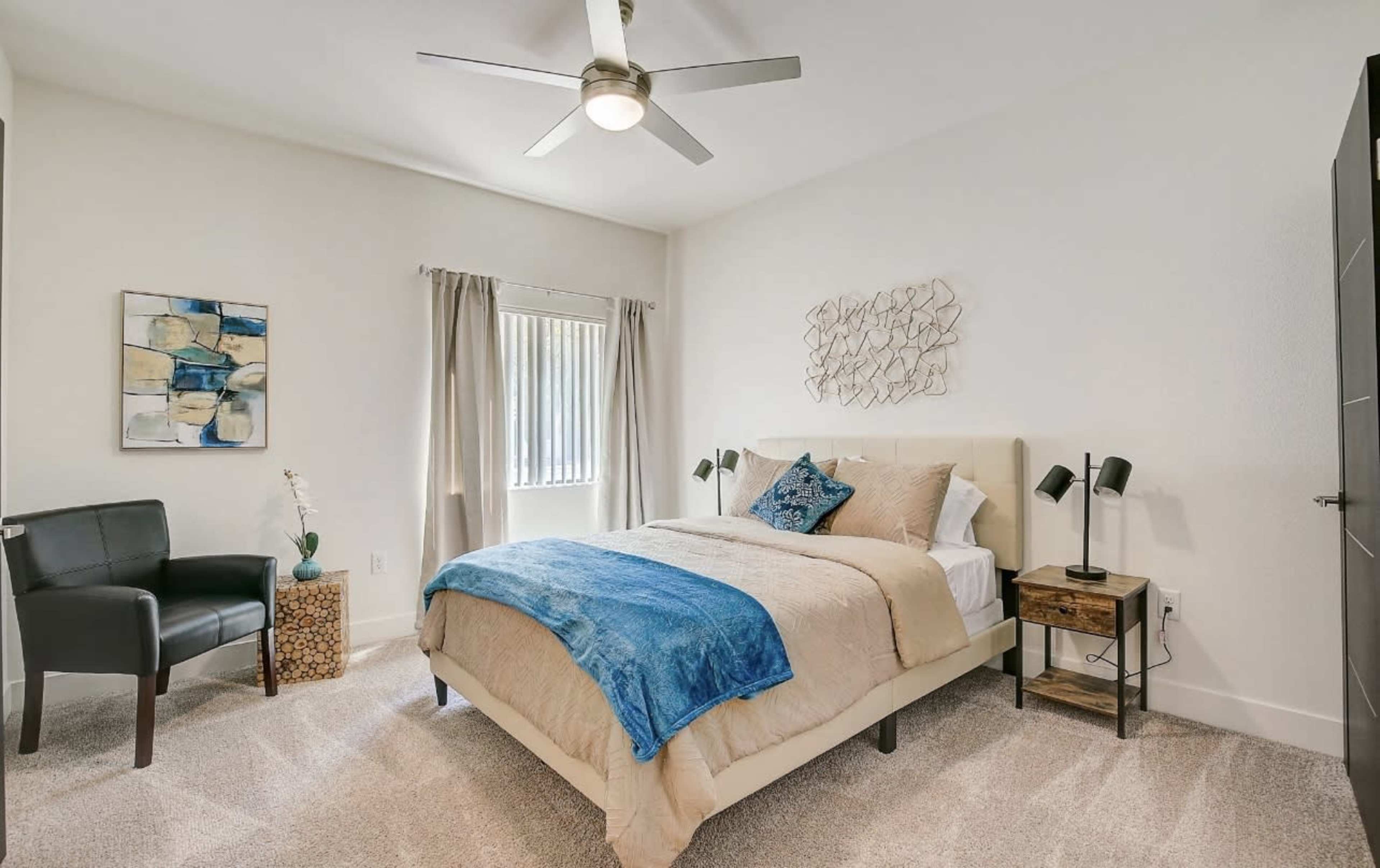 A bedroom with a bed, a chair, and a ceiling fan, featuring light-colored walls and a window with curtains.