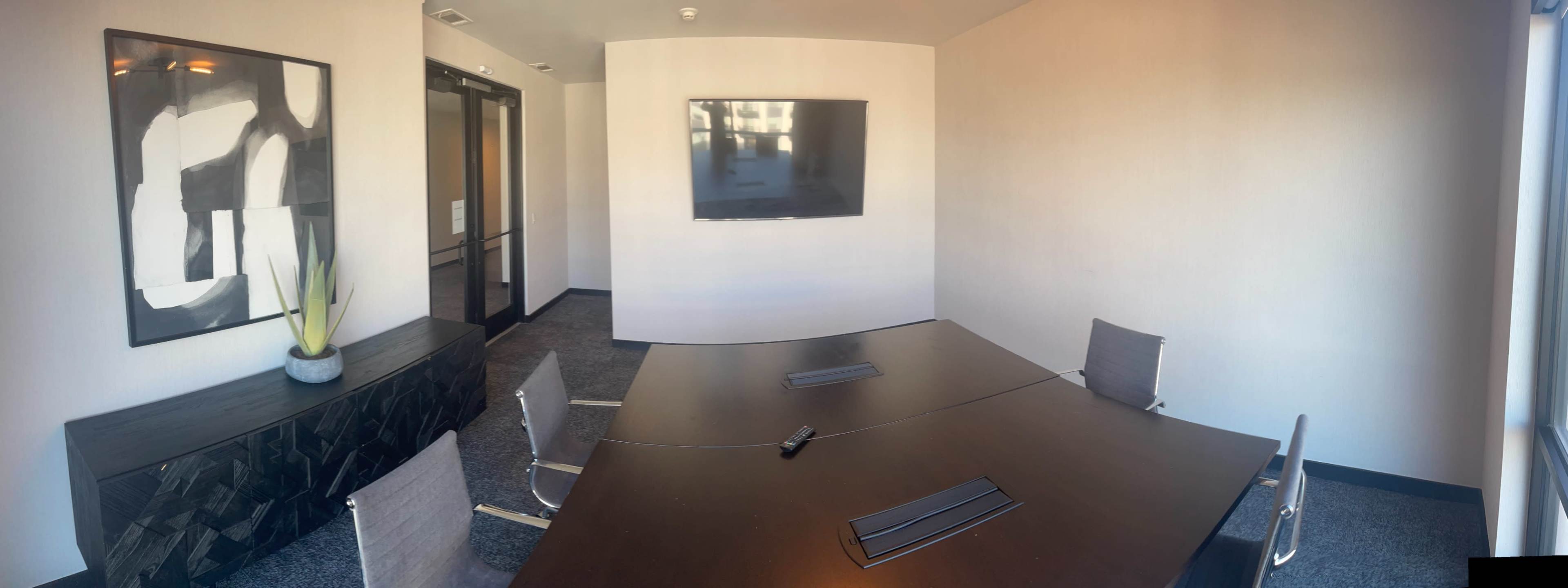 The image shows a modern conference room with a large table, gray chairs, a television mounted on the wall, and minimal decorations.
