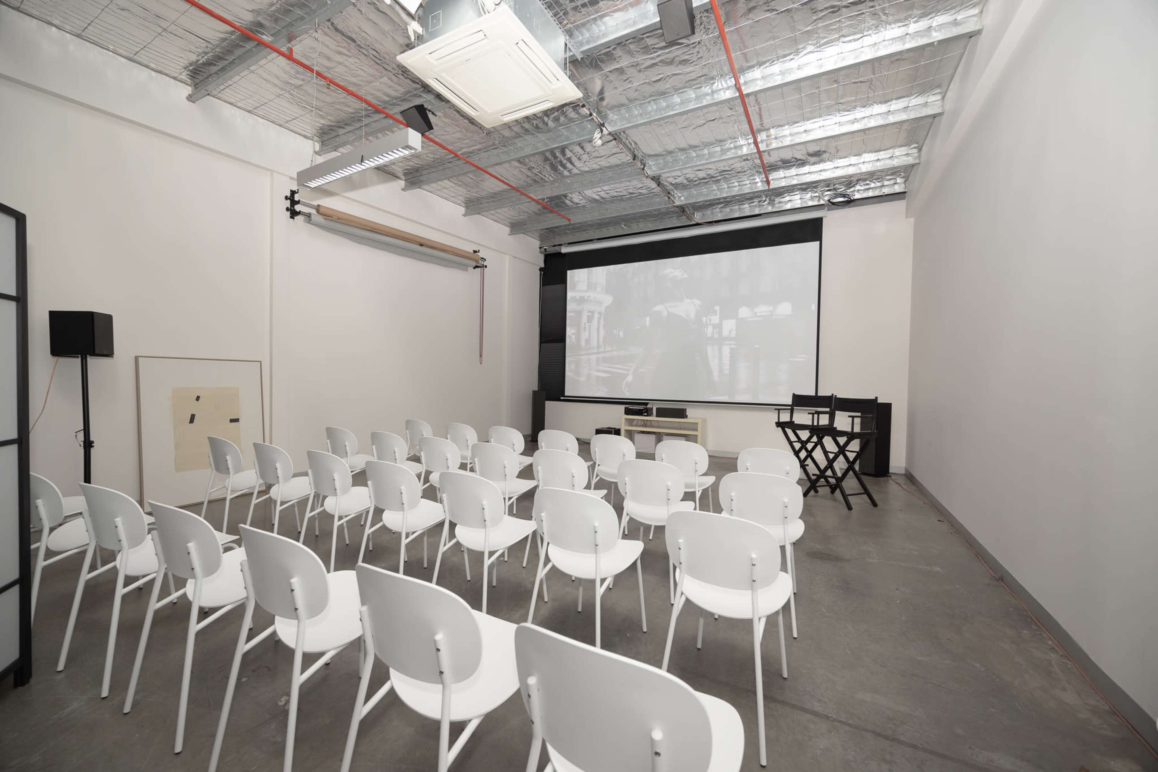 The image shows an empty presentation room with rows of white chairs facing a large screen on the wall.