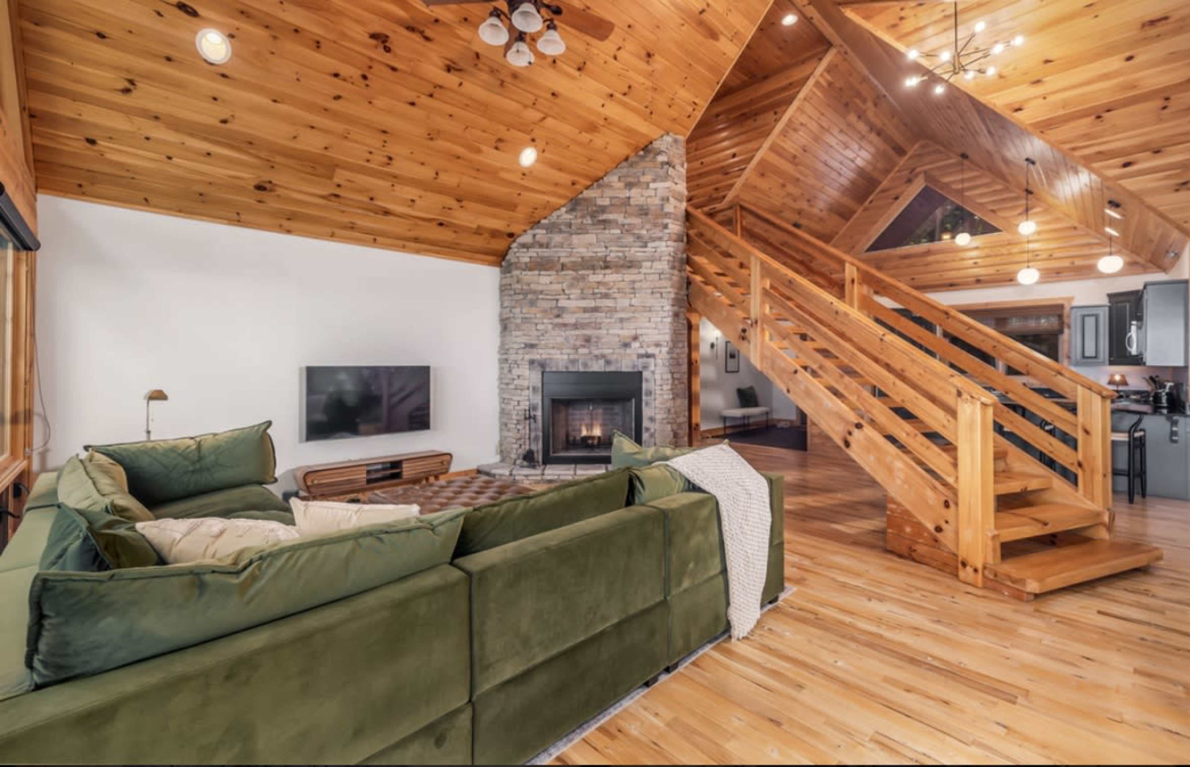 A spacious living area features a large green sectional sofa, a stone fireplace, wooden beams, and an open staircase leading to the upper floor.