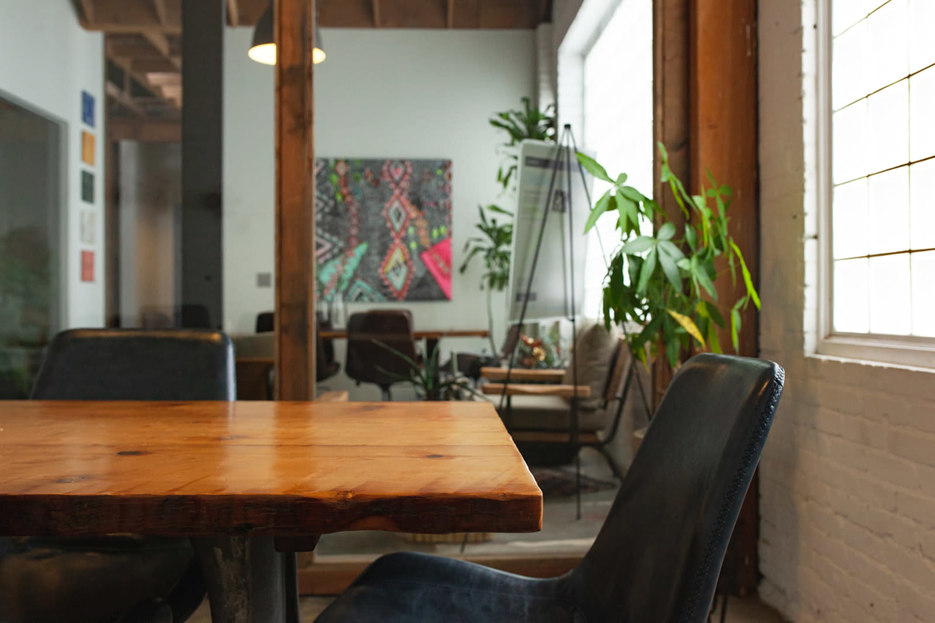 A wooden table with a natural finish is positioned in the foreground, while a modern workspace with plants and artwork is visible in the background.