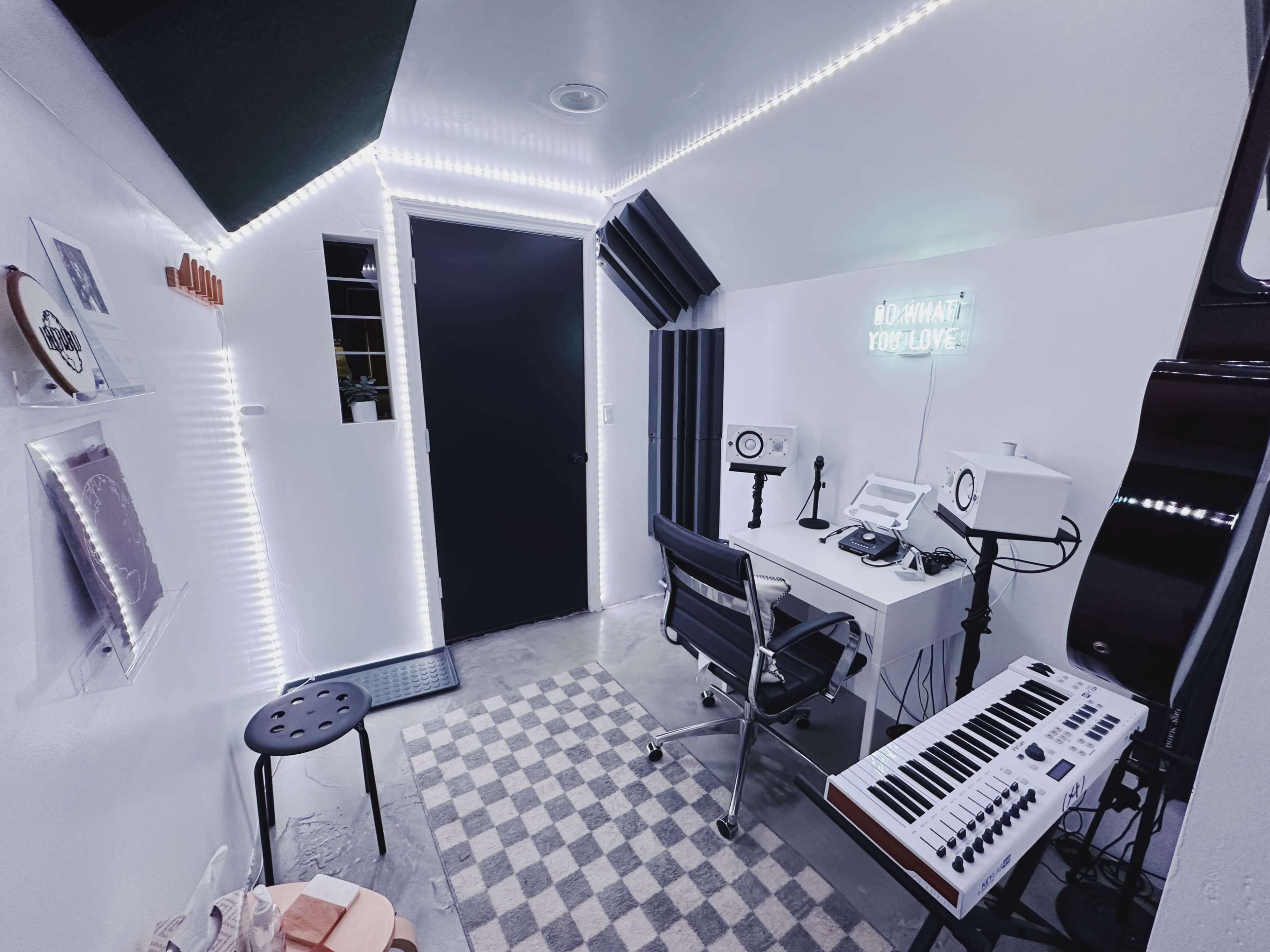 A small music studio featuring a desk with equipment, a keyboard, and acoustic panels, all illuminated by LED lights along the walls.