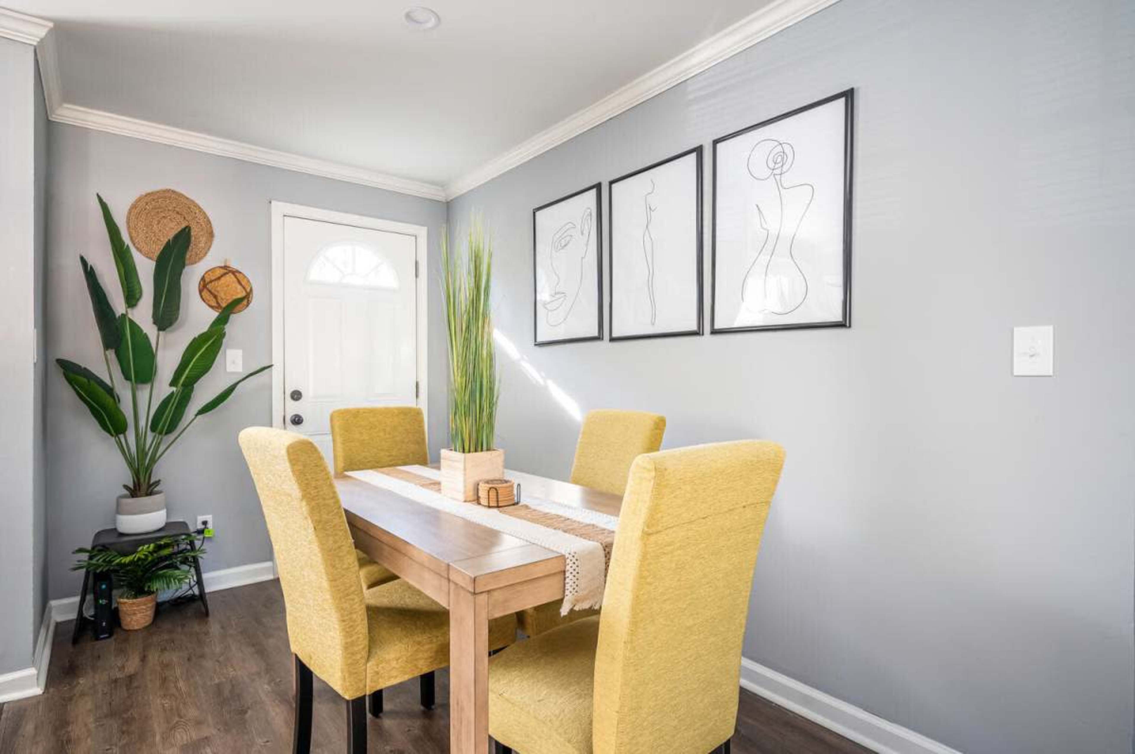 A dining area features a wooden table surrounded by four yellow chairs, with a large green plant in one corner and framed line art on the wall.