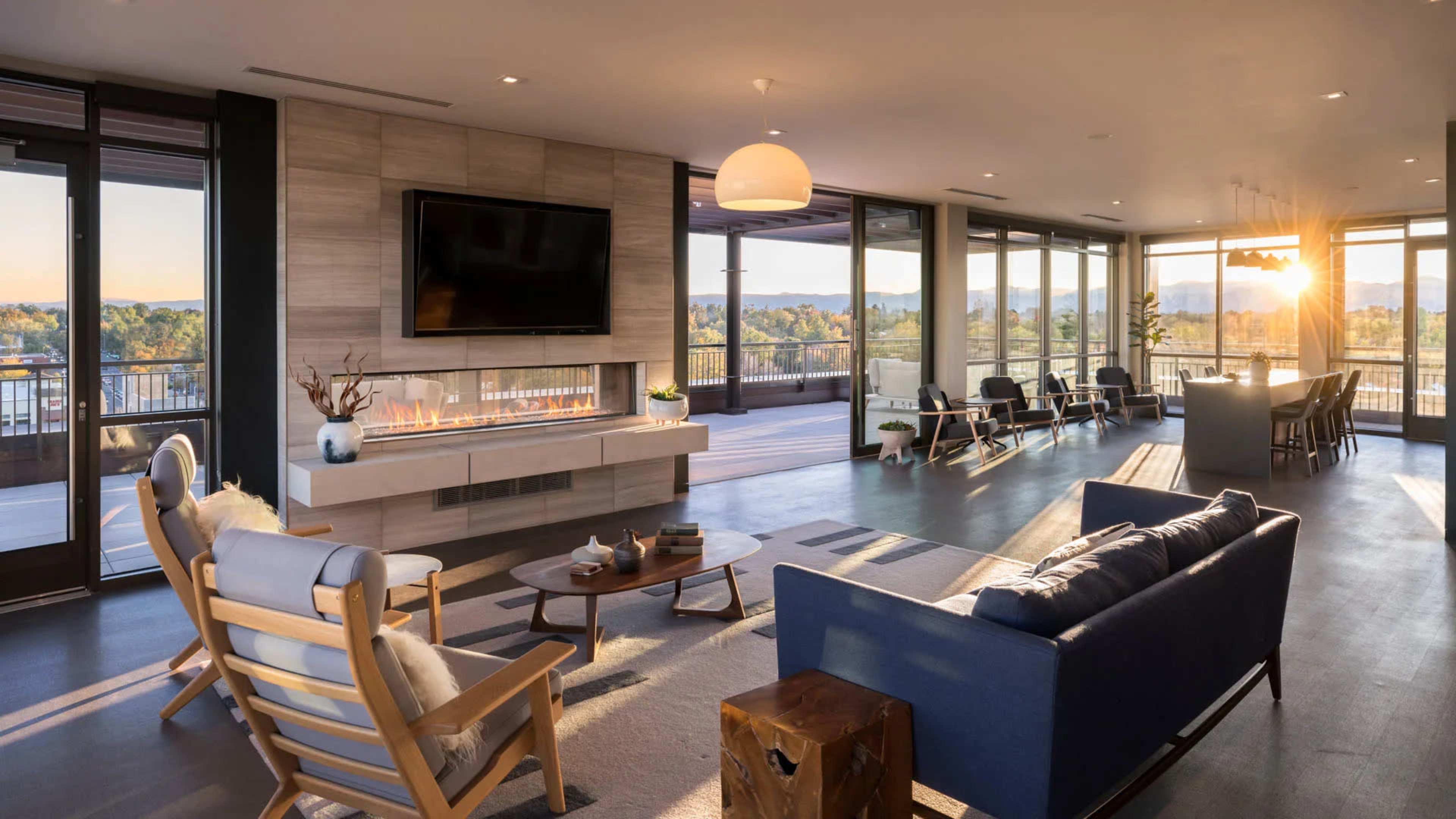 A modern living room with large windows, a television mounted on the wall, a fireplace, and contemporary furniture, including a wooden coffee table and lounge chairs.