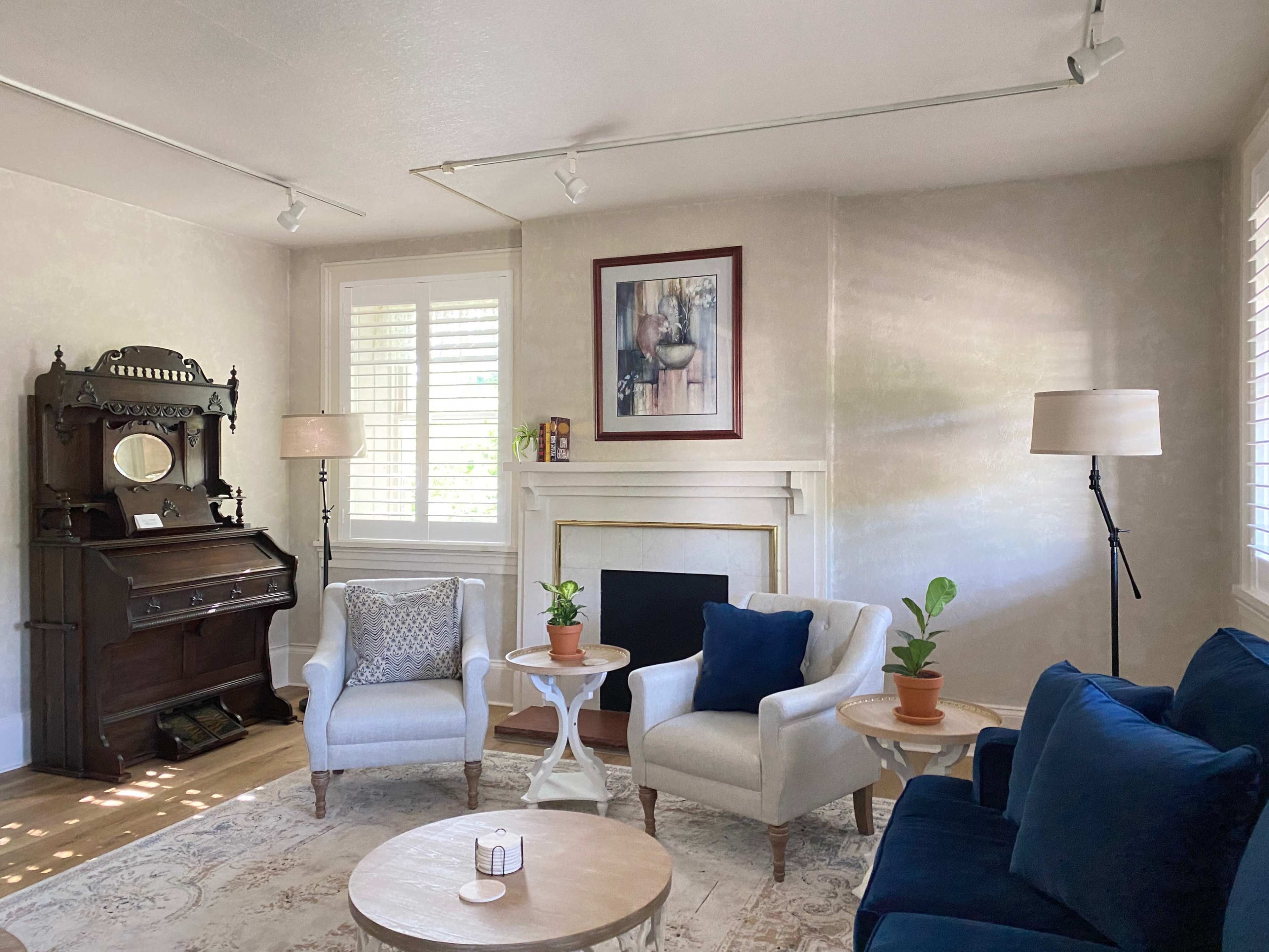 A cozy living room with a vintage piano, two armchairs, a coffee table, and indoor plants arranged around a fireplace.